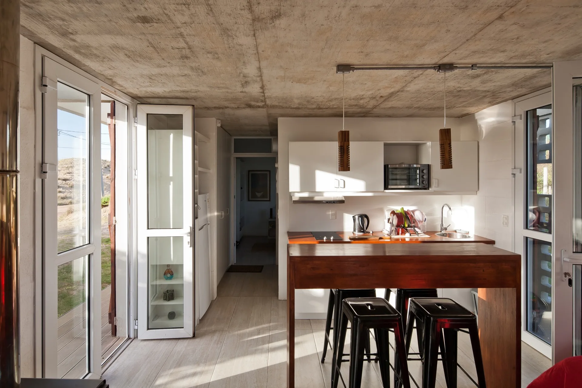 A modern kitchen features an exposed concrete ceiling, white cabinetry, a warm wood island with black metal stools, and glass doors leading outside.