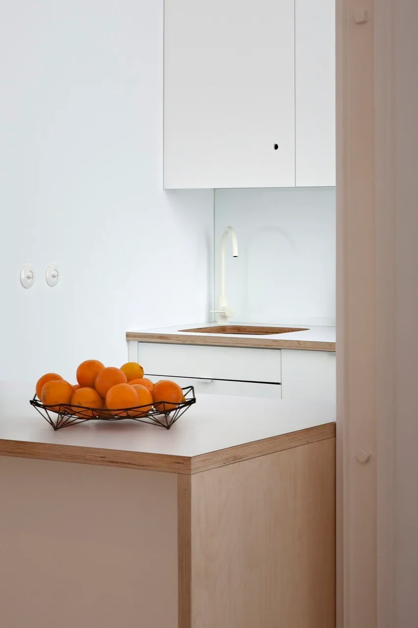 A minimalist white kitchen featuring handleless cabinets, a white gooseneck faucet, and a plywood-edged island with a bowl of oranges.