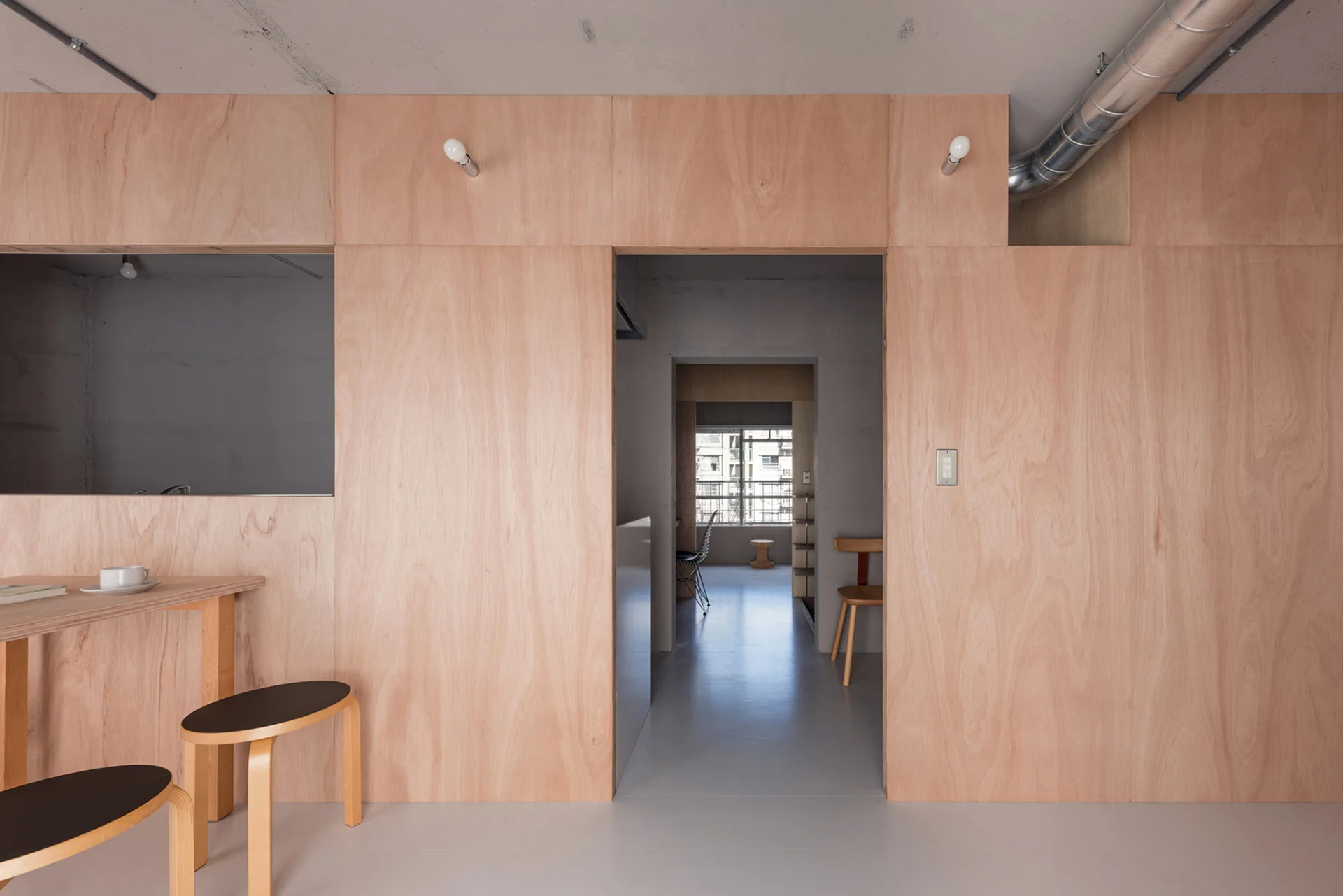 A minimalist apartment interior features light wood plywood wall panels, a grey concrete ceiling with exposed ductwork, and wooden dining furniture.