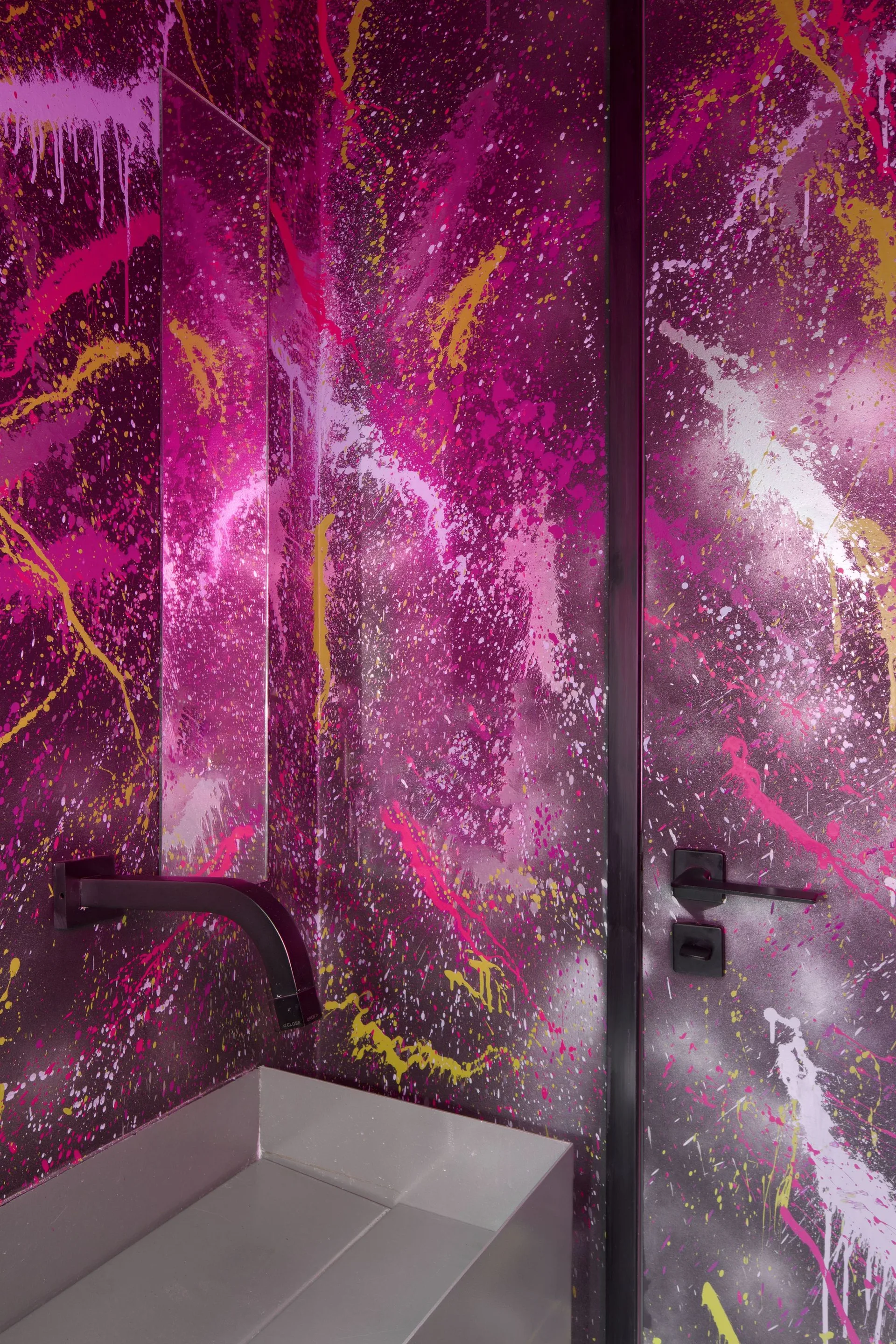 A modern bathroom features vibrant magenta paint splatter walls, a rectangular metal sink, and matte black fixtures including a wall-mounted faucet.