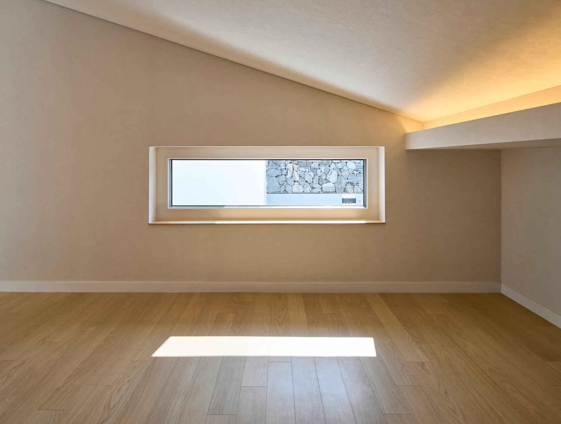An empty minimalist attic room featuring a sloped ceiling, light wood floors, indirect cove lighting, and a low horizontal window.