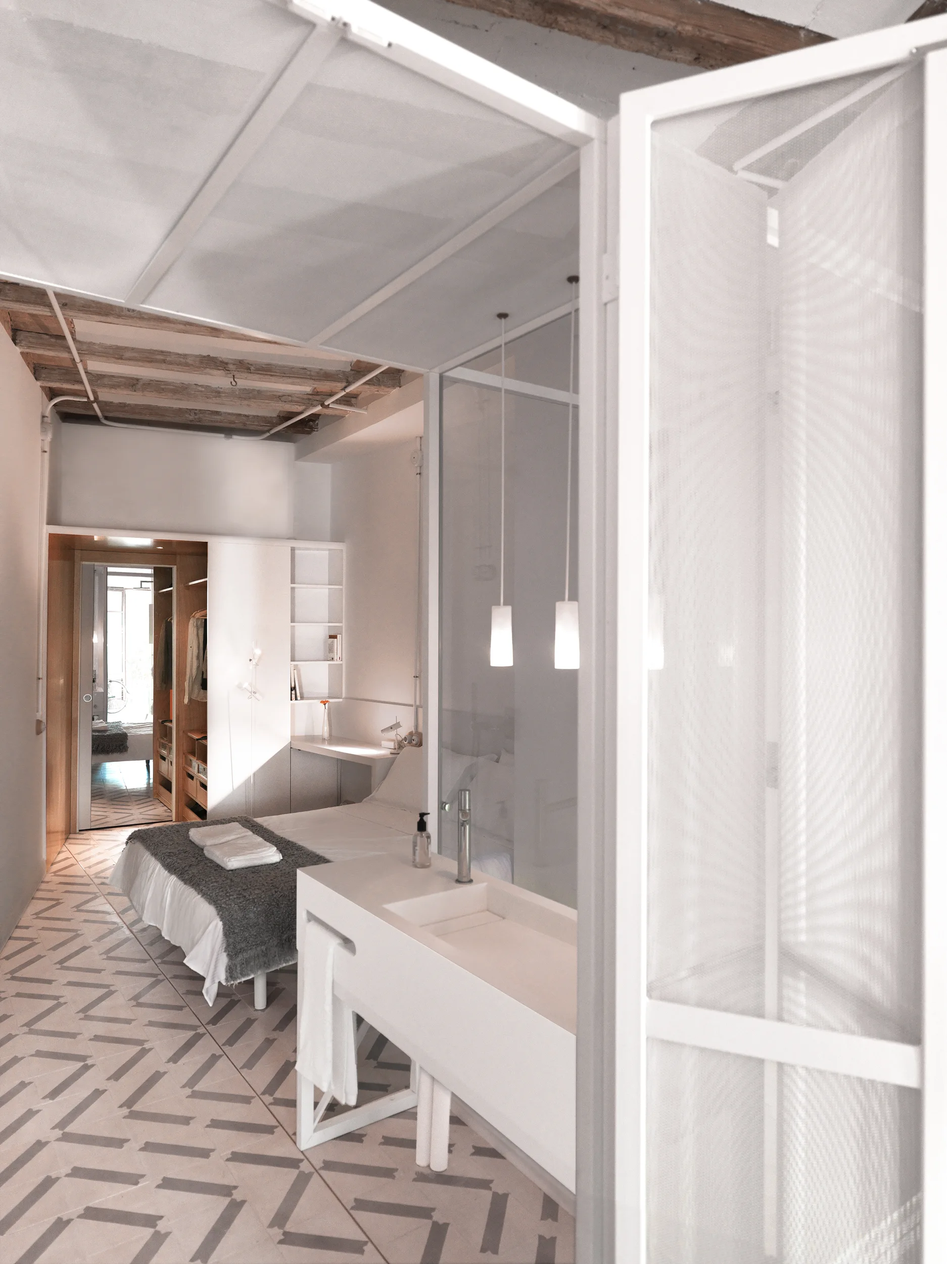 An open-concept bedroom features a white vanity, a bed with grey throw, patterned floor tiles, and exposed wood ceiling beams.