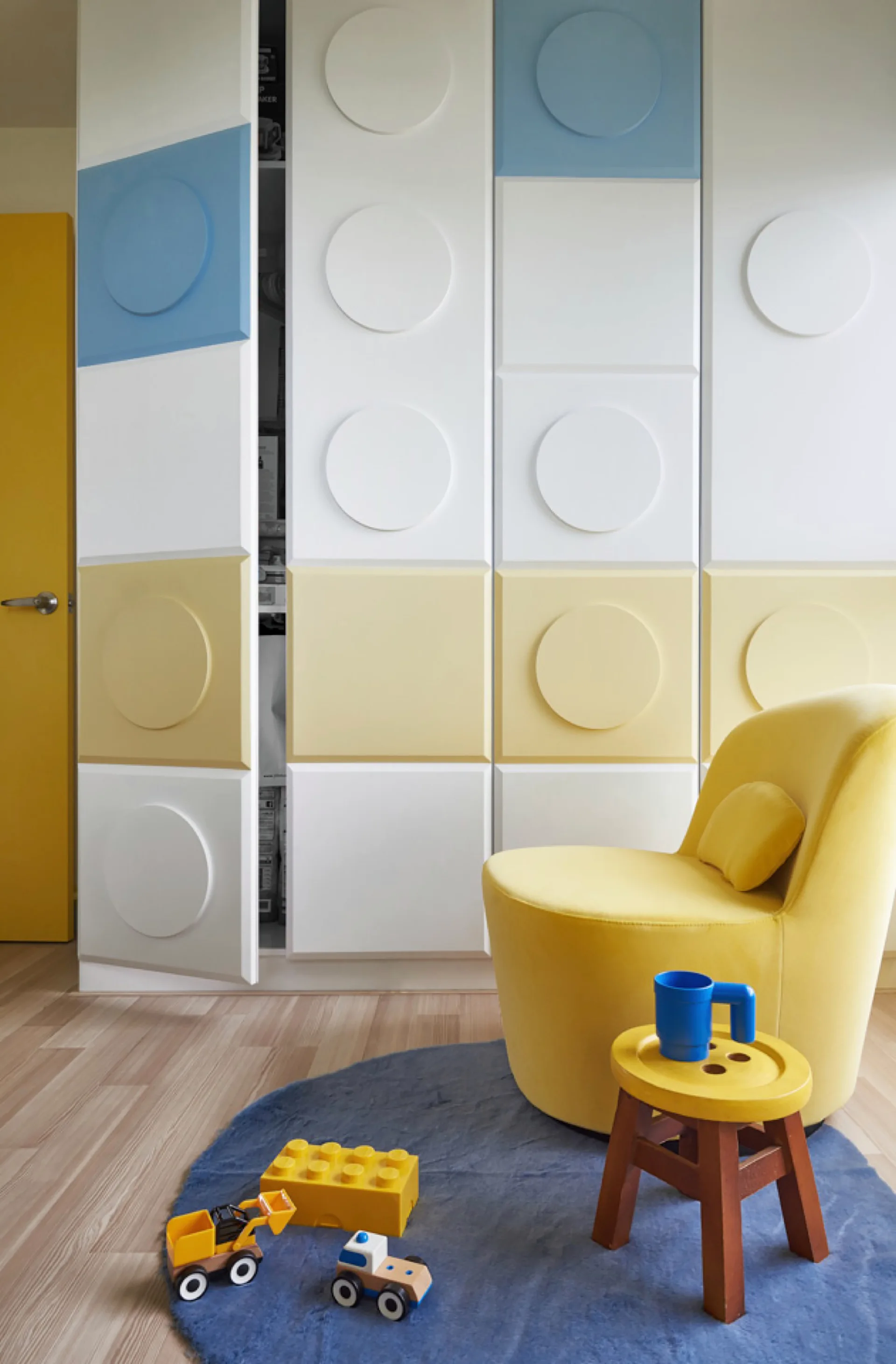 A children's room features a yellow upholstered armchair next to a white, yellow, and blue Lego-themed storage wardrobe and a blue circular rug.