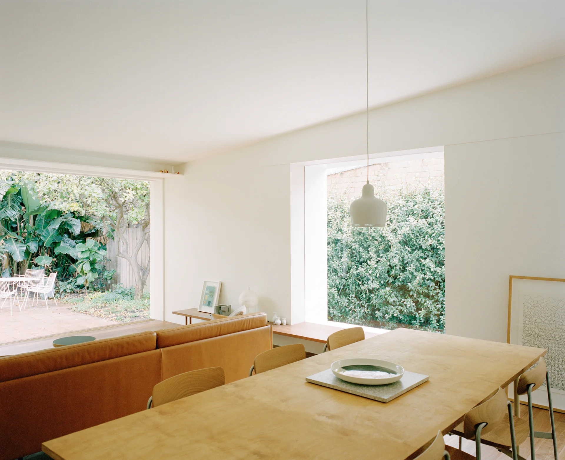 A light-filled dining space features a long wooden table, a tan leather sofa, and a large window showcasing garden greenery.