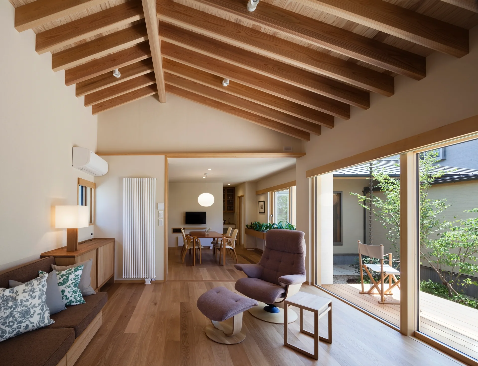 A minimalist living room with a vaulted wooden ceiling, an armchair with an ottoman, and glass sliding doors opening to an outdoor deck.