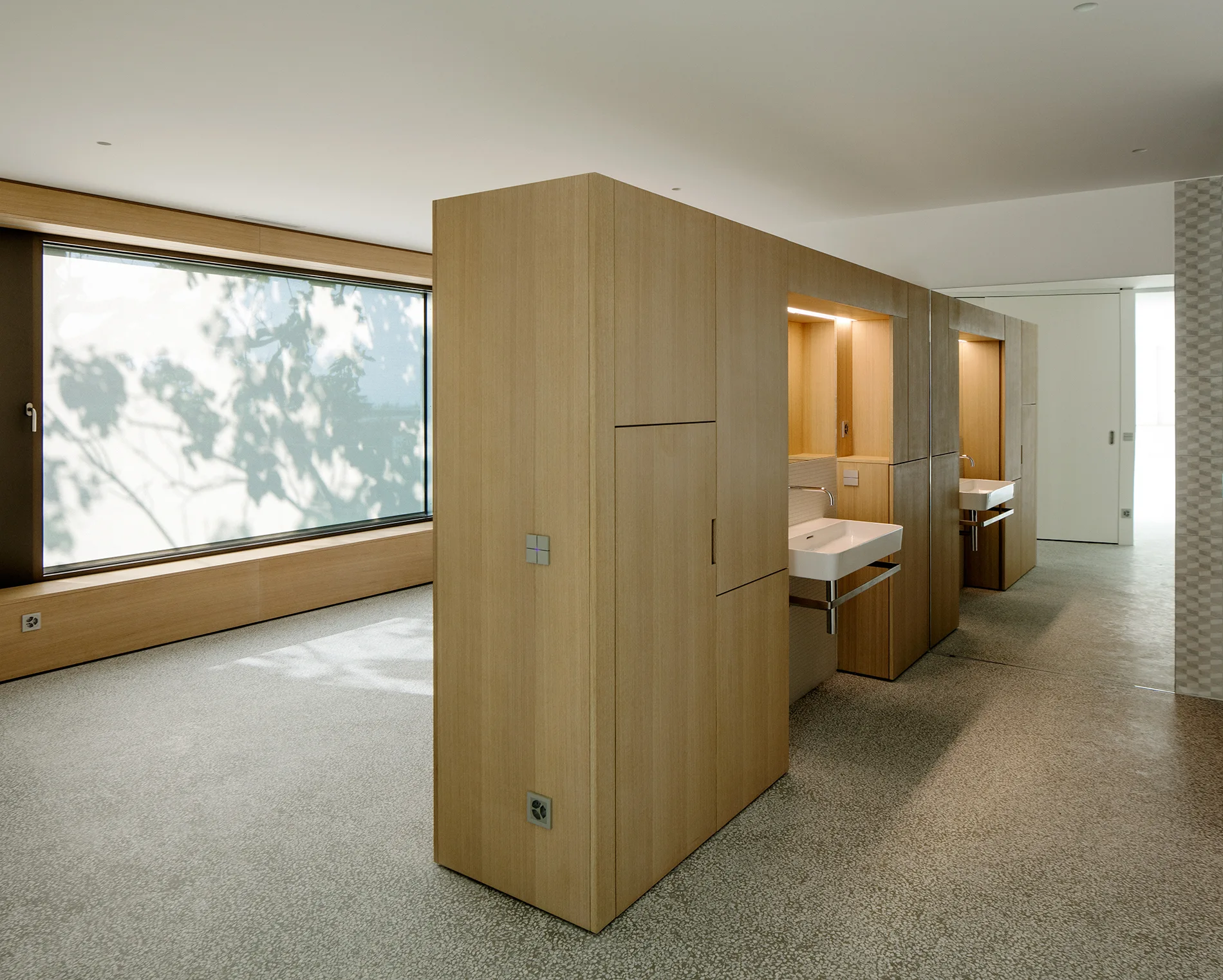 A minimalist bathroom featuring a large freestanding light wood cabinet unit with integrated sinks, set against a low wood bench and speckled terrazzo flooring.
