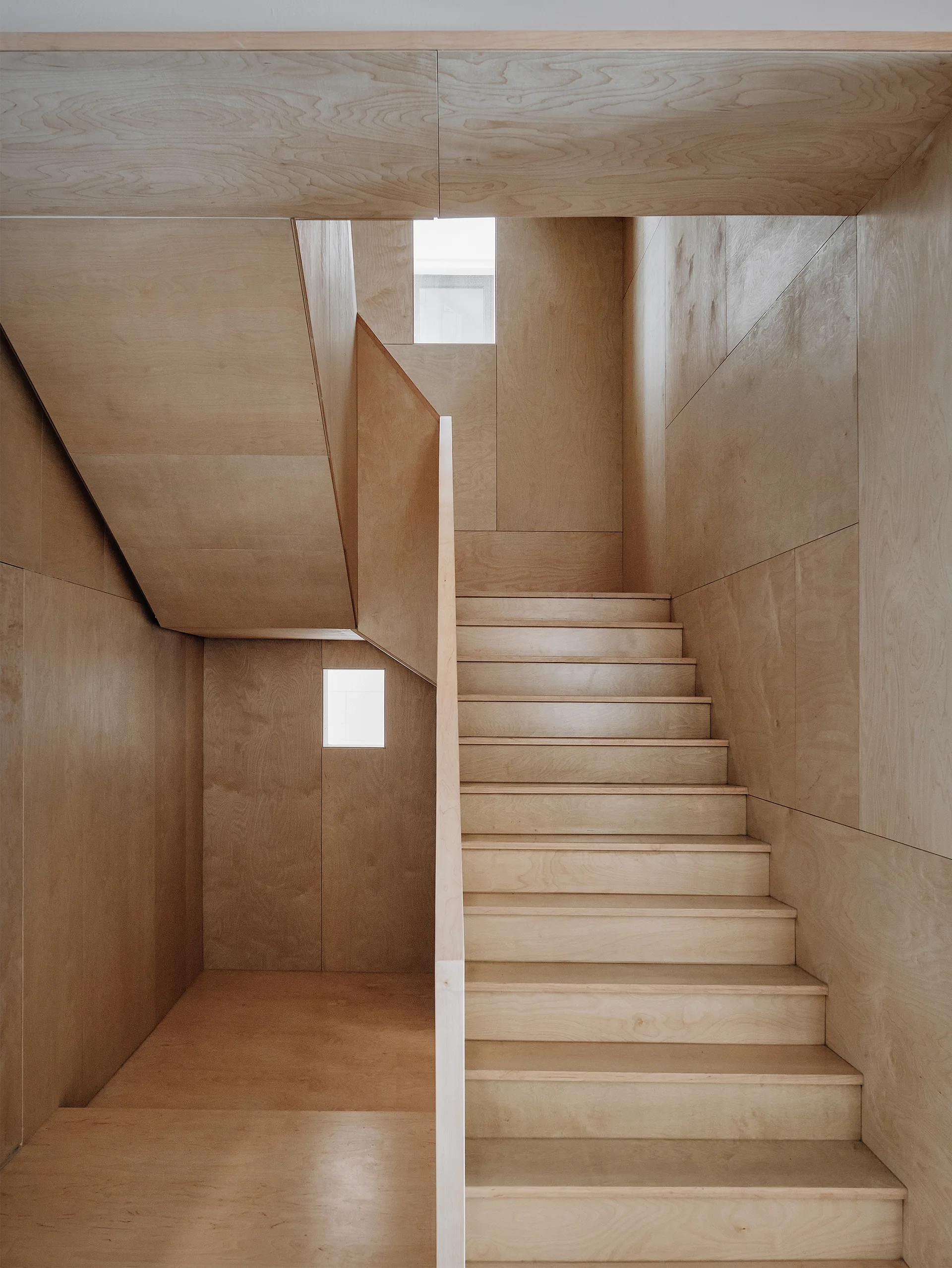 A minimalist wooden staircase and surrounding walls constructed entirely from light-toned plywood panels with two small square windows.