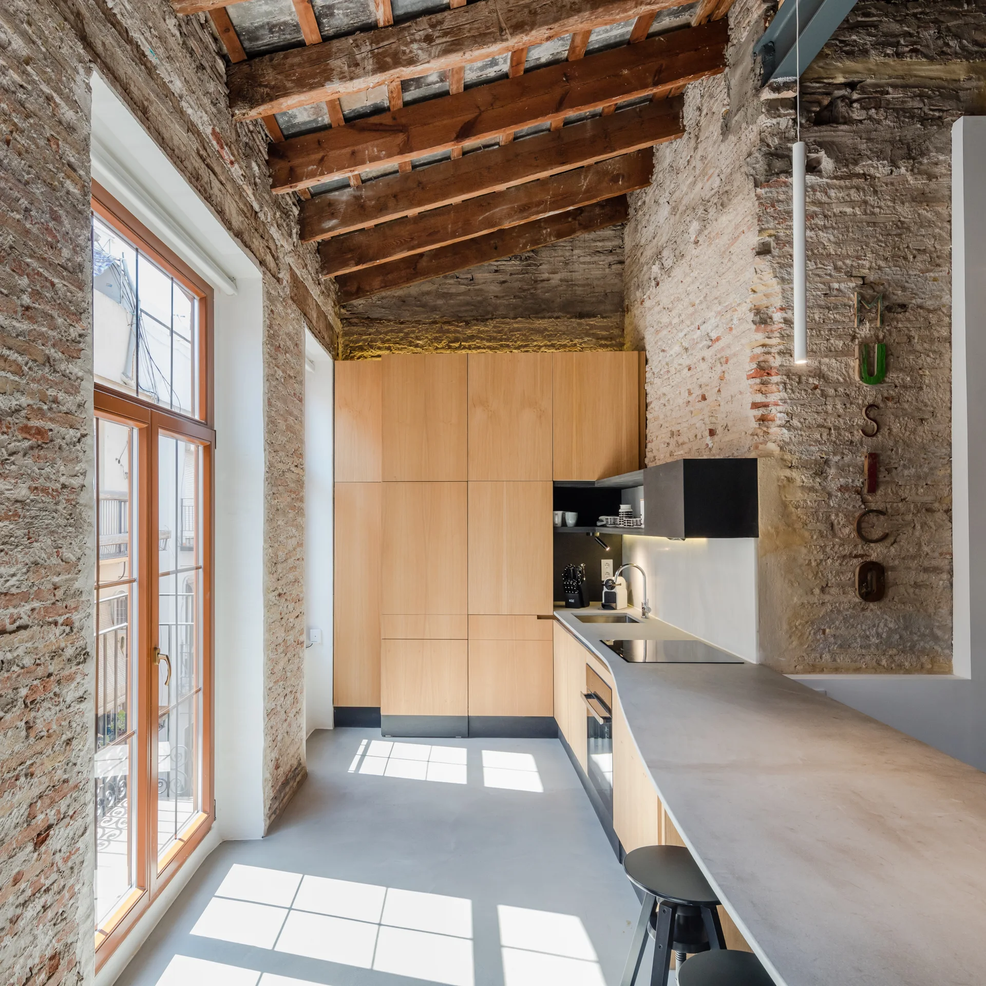 A modern loft kitchen features flat-panel light wood cabinets, a long grey countertop, exposed brick walls, and vaulted wooden ceiling beams.