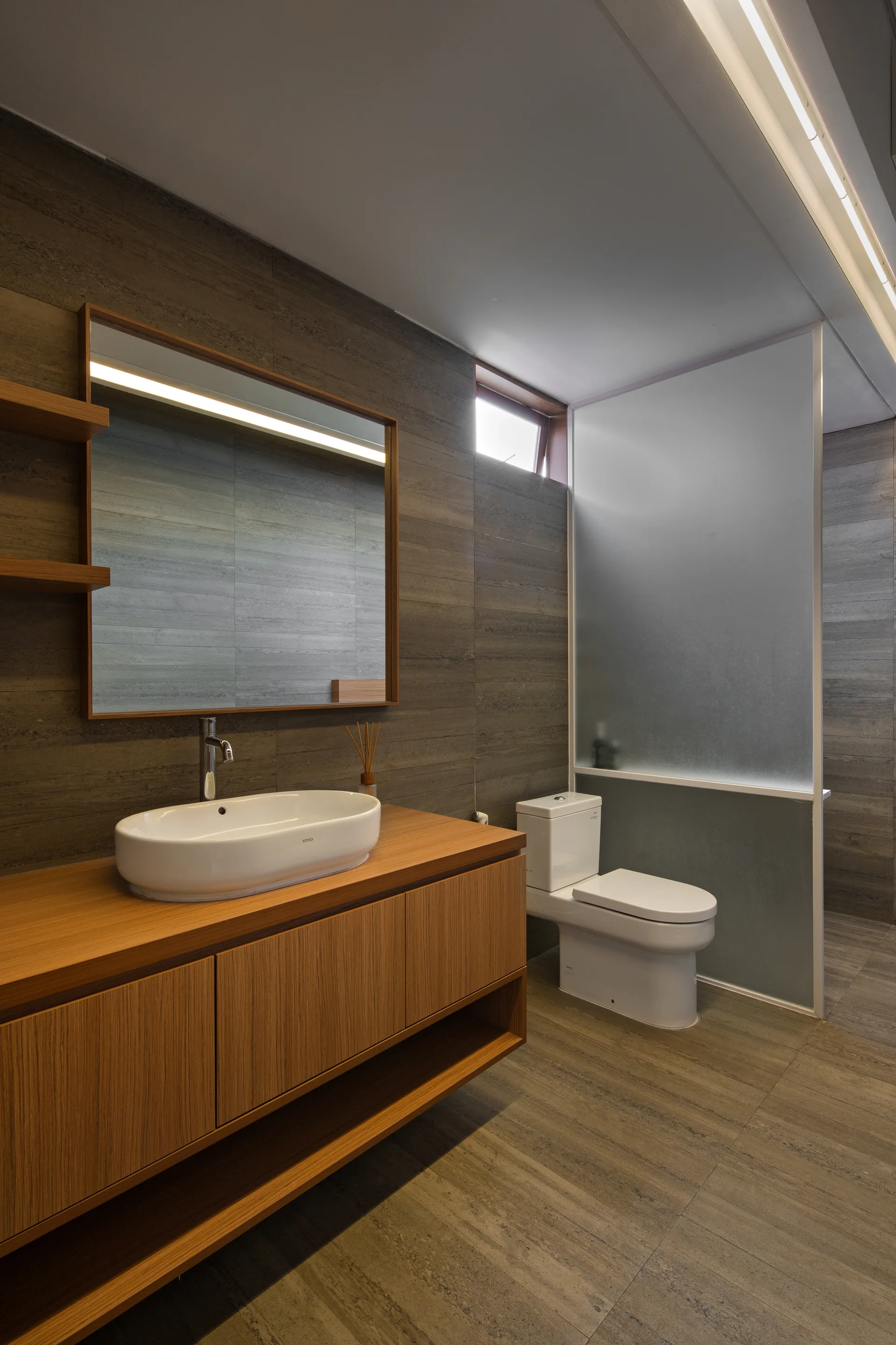 A modern bathroom features a wooden floating vanity with a white vessel sink, a white toilet, and textured brown stone wall tiles.