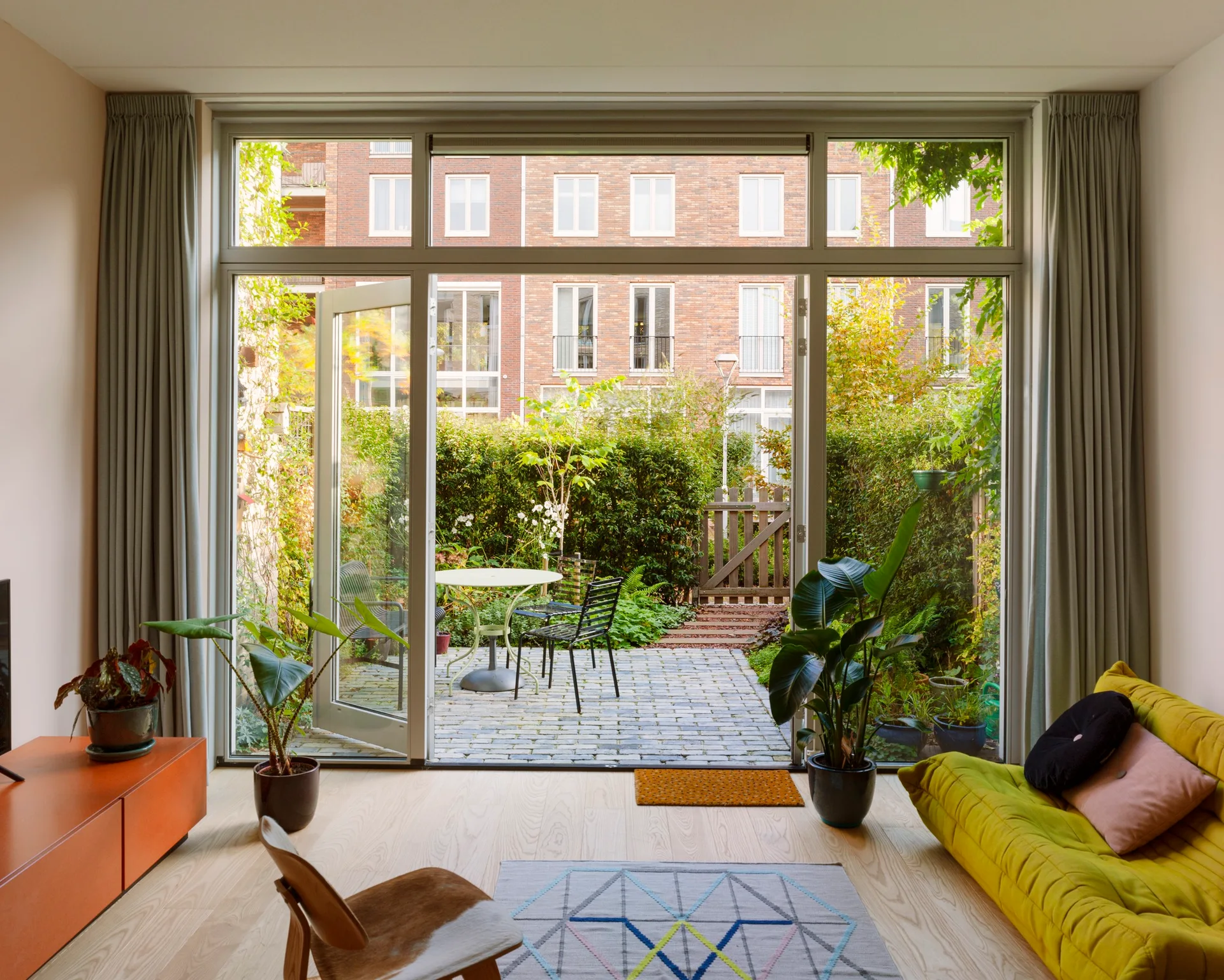 A bright living room with a yellow tufted sofa and light wood floors opening to a paved patio garden with a table and chairs.