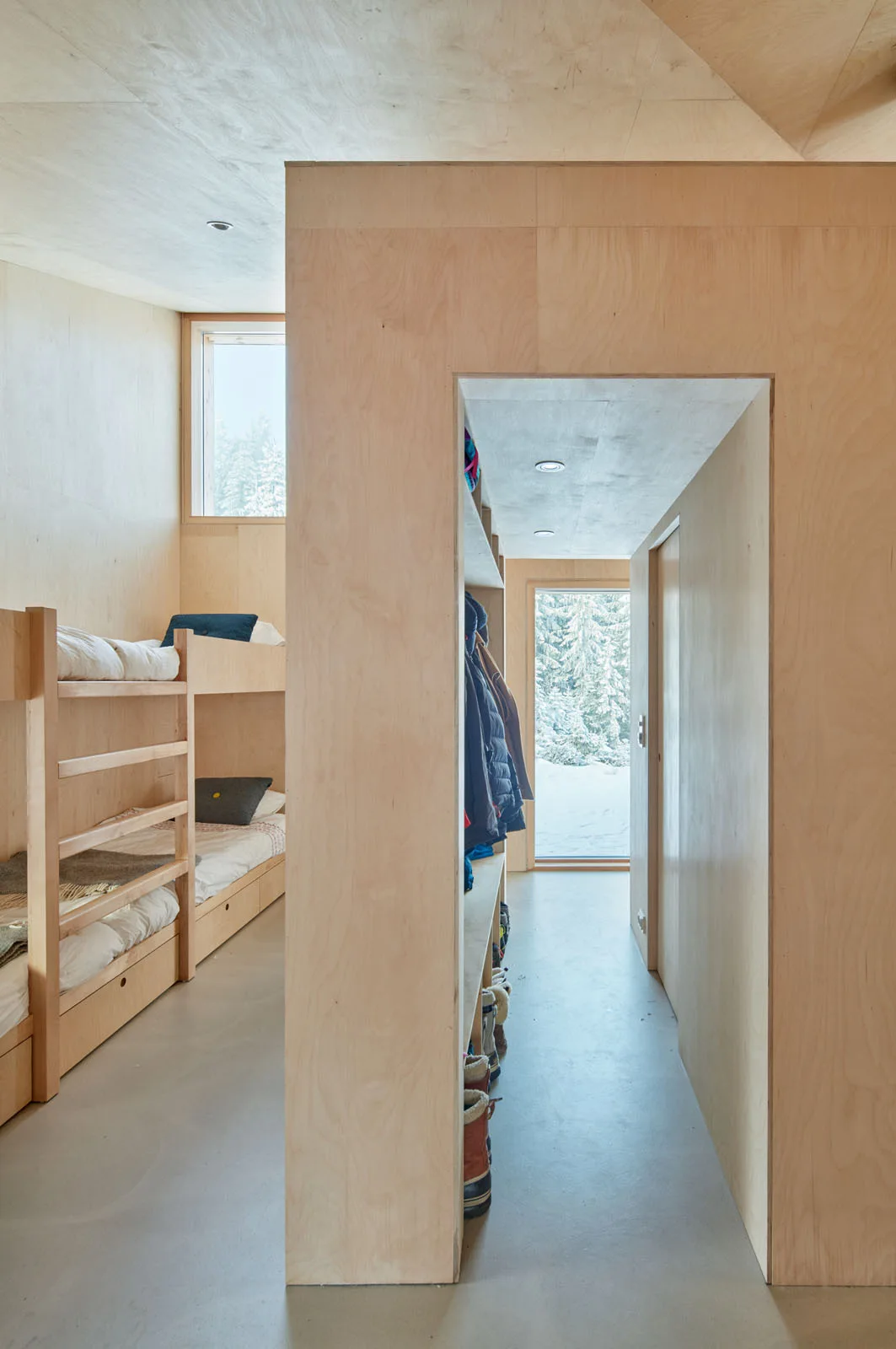 A minimalist cabin bedroom features light plywood walls, built-in wooden bunk beds, and a partition wall leading to an entryway with coat storage.