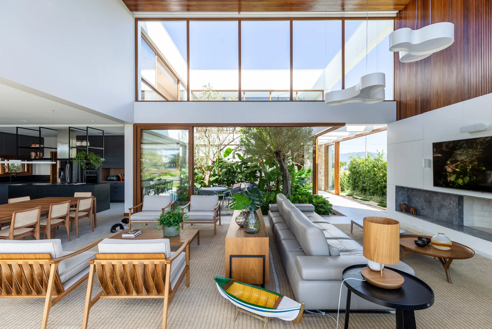 A spacious open-plan living room with wood armchairs and a gray leather sofa features large glass walls looking onto an indoor garden courtyard.