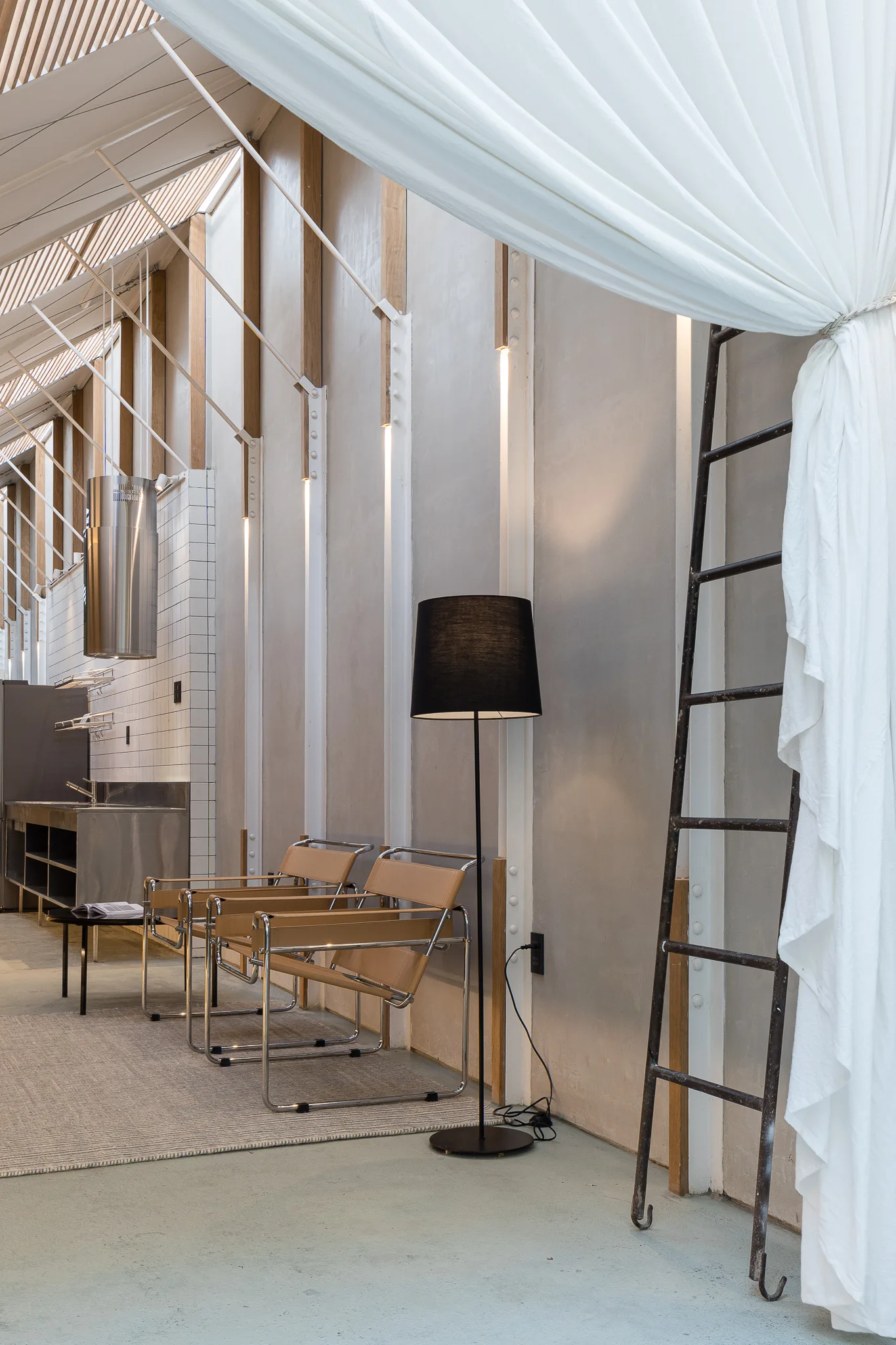 Two tan leather sling chairs are arranged next to a black floor lamp and a metal ladder in a high-ceilinged room with vertical wall lights.