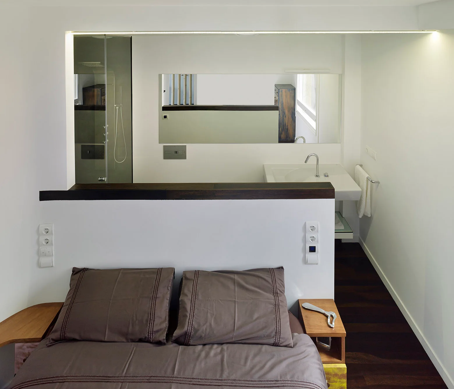 A modern bedroom features a low white partition wall separating the brown-dressed bed from an integrated bathroom with a vanity and mirror.