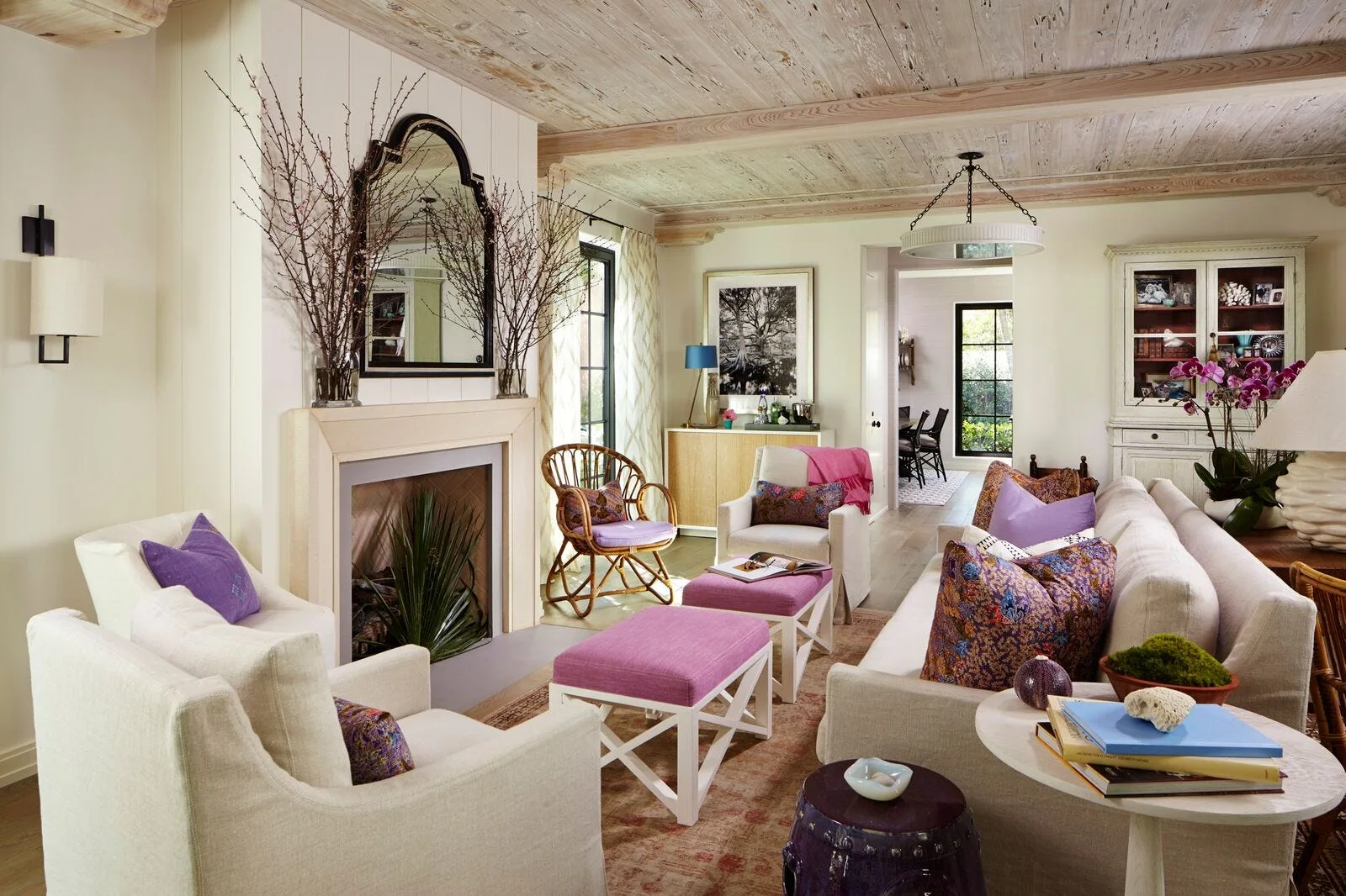 A bright living room features a stone fireplace with an arched mirror, cream sofas, purple ottomans, and a rustic wood plank ceiling.