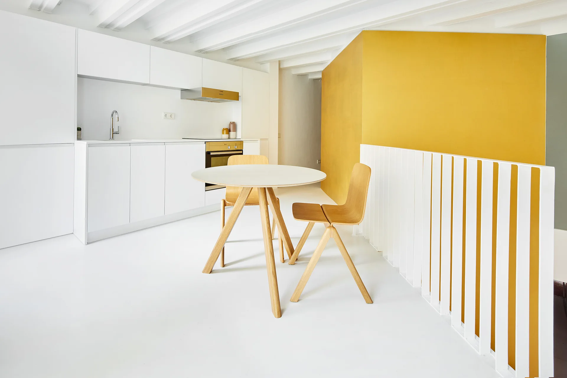 A minimalist white kitchen featuring a round wooden dining table, white ceiling beams, and a bold yellow accent wall next to a vertical slat railing.