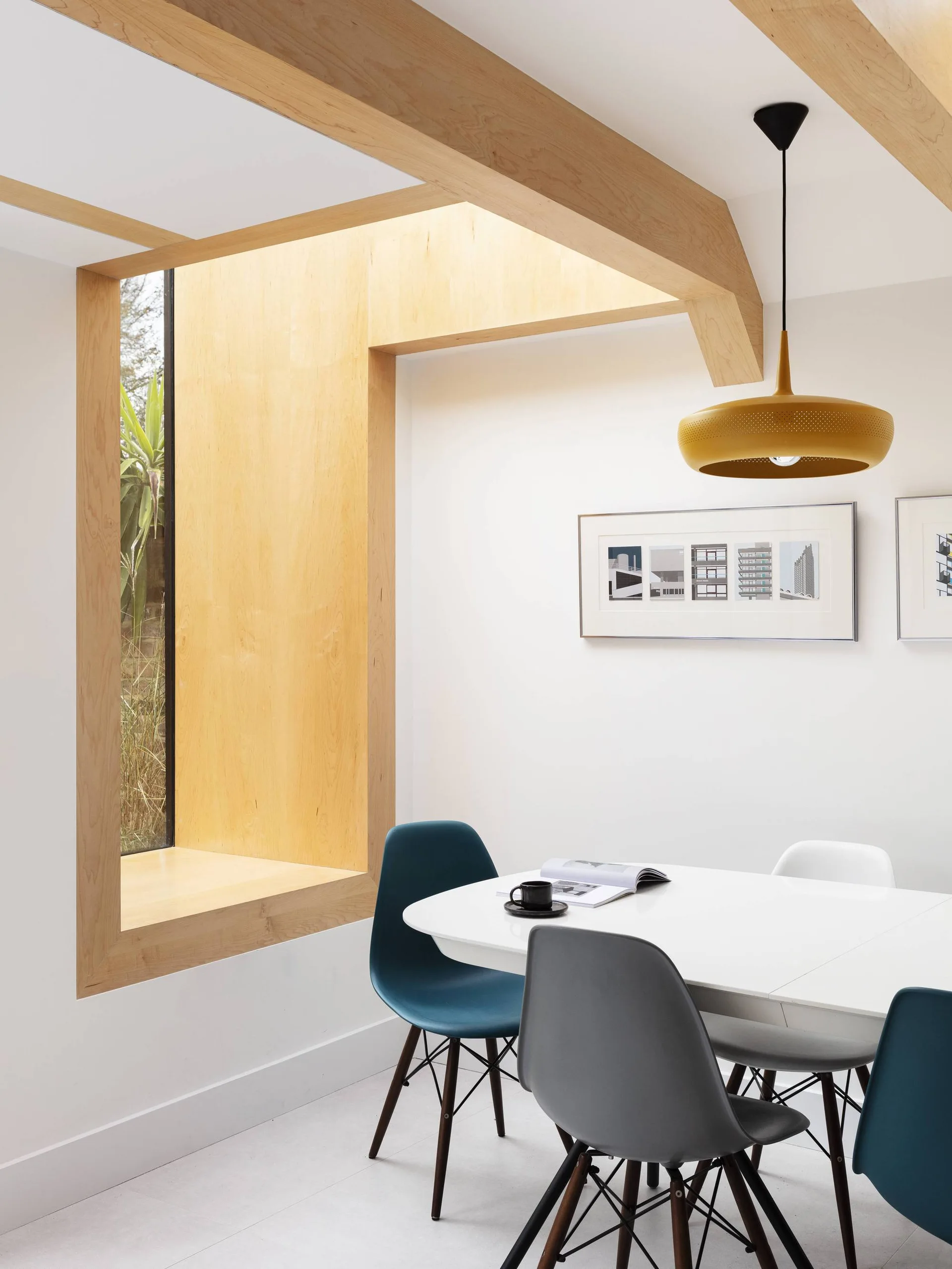 A modern dining area featuring a white table with blue and grey molded chairs, a light wood-lined window nook, and a yellow pendant light.