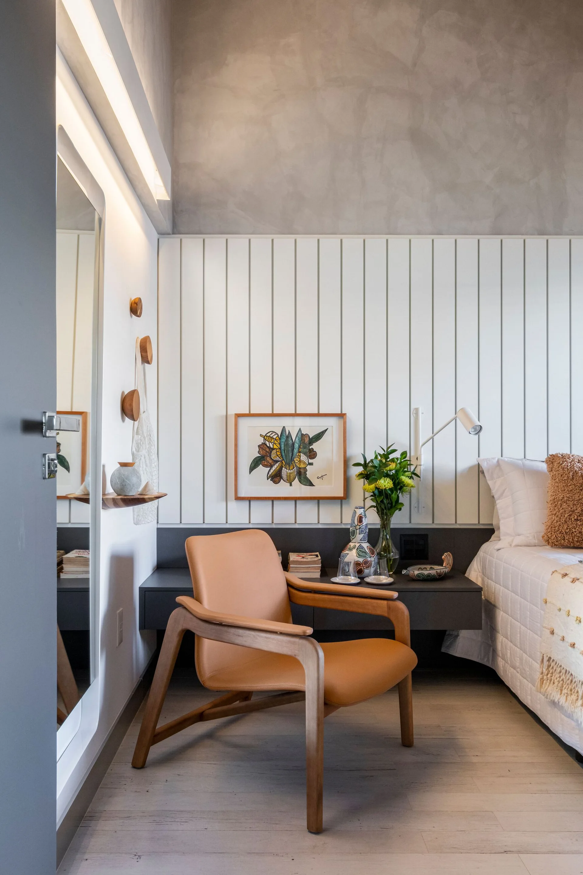 A modern bedroom corner featuring a tan leather armchair in front of a white slatted wall panel, with a dark grey floating bedside drawer.