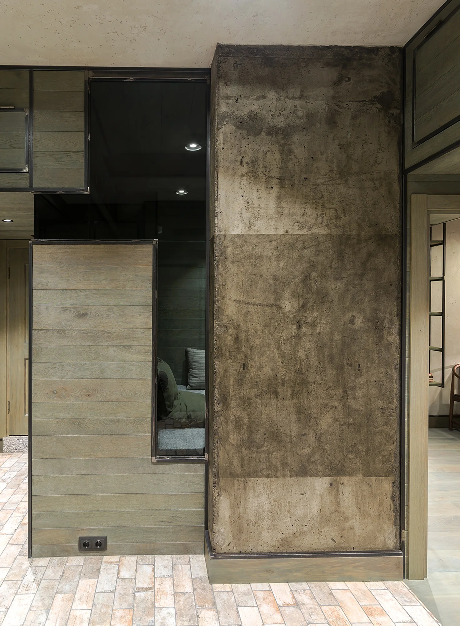 An interior partition wall featuring muted green-grey wood paneling with black metal trim adjacent to a textured, raw concrete column.