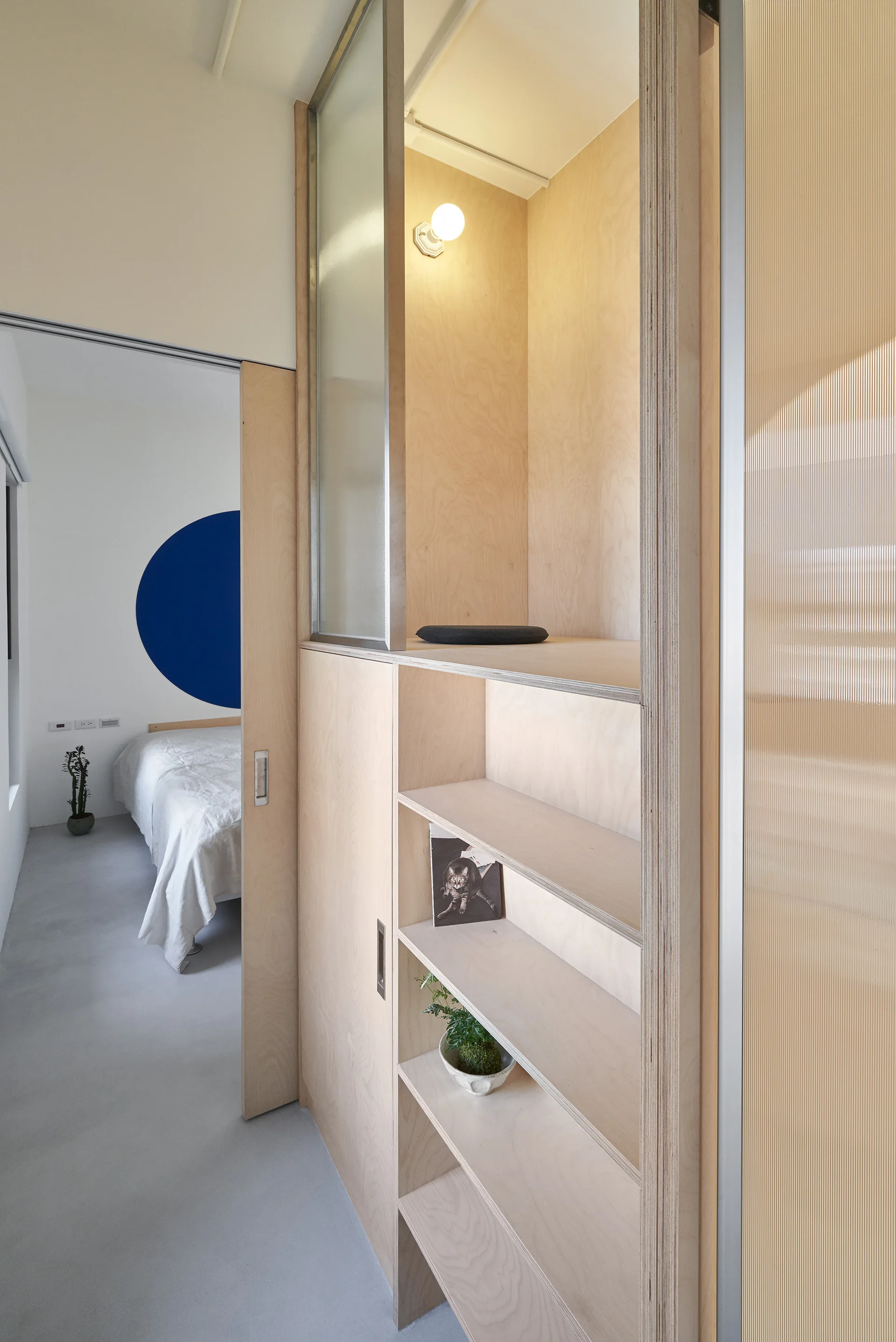 A minimalist bedroom featuring a custom plywood built-in shelving unit, a sliding wooden door, and a blue circular graphic on the back wall.