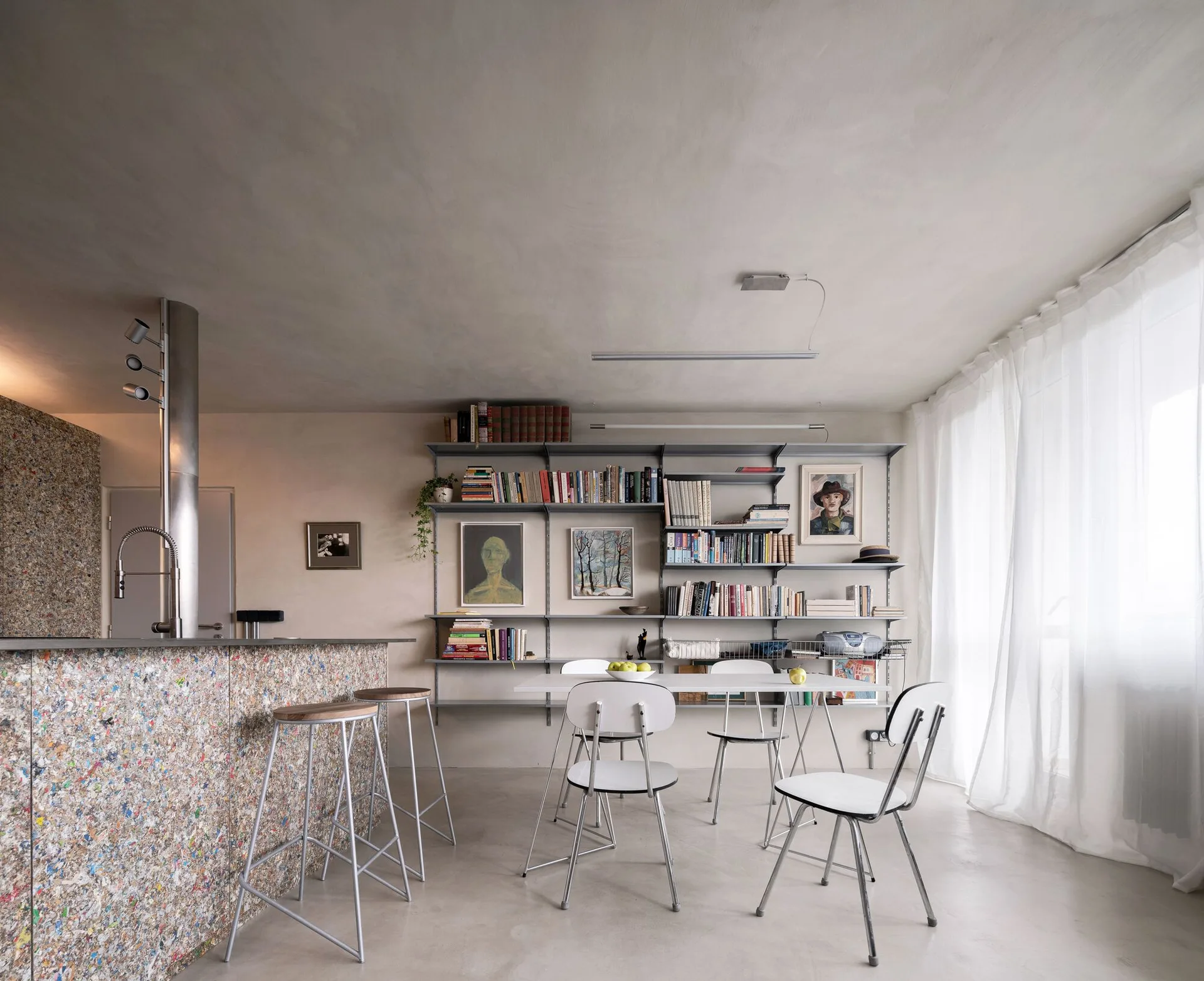 A minimalist dining room featuring a white dining set, modular metal wall shelving, and a kitchen island with a speckled multicolor facade.