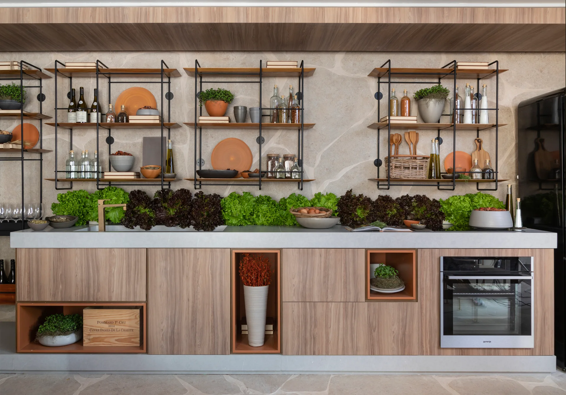 A modern kitchen with wood-toned cabinets, black metal open shelving, a grey countertop lined with green lettuce, and an irregular stone wall.