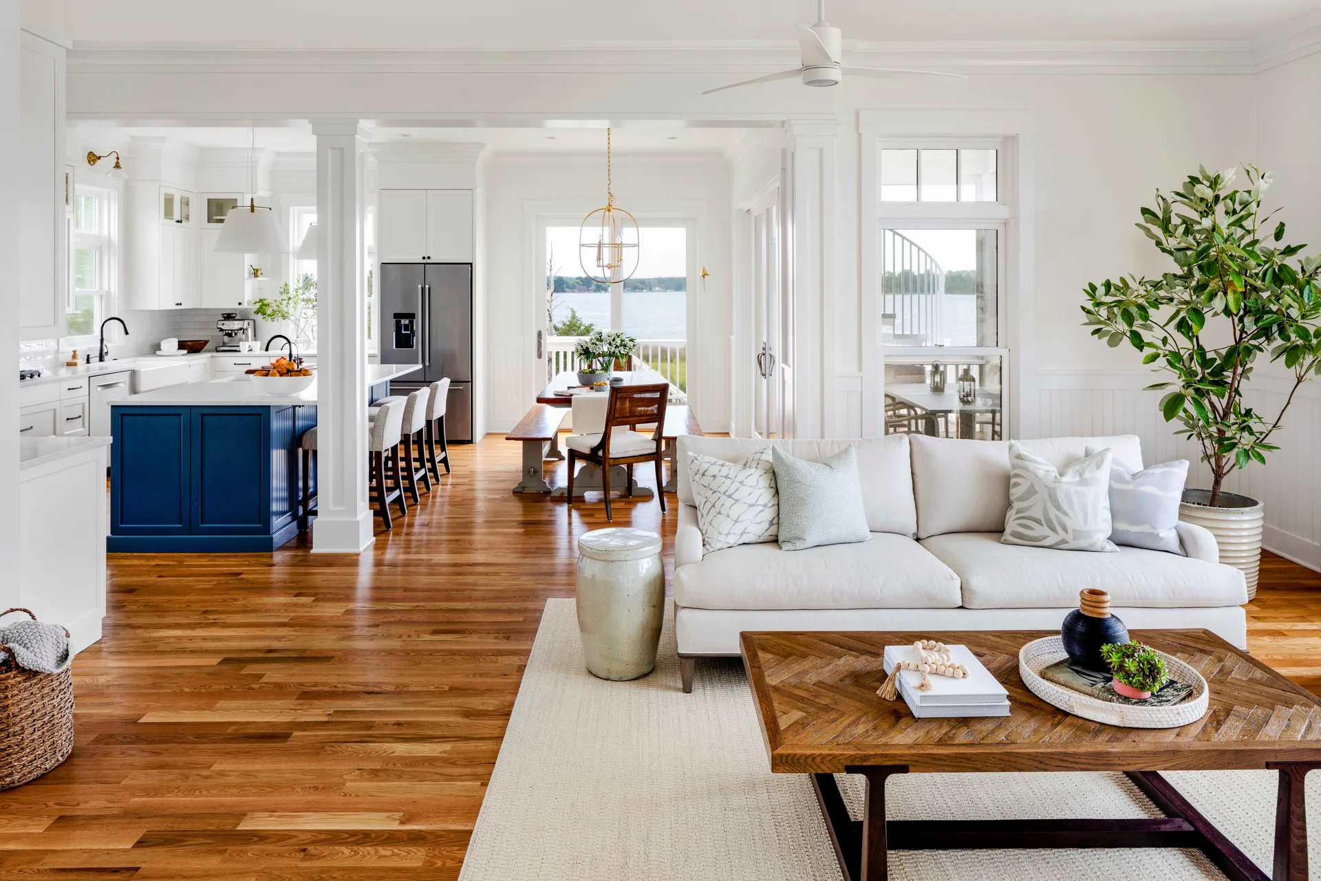 An open-concept living area features a cream sofa, a herringbone wood coffee table, and a kitchen with a navy blue island against white walls.