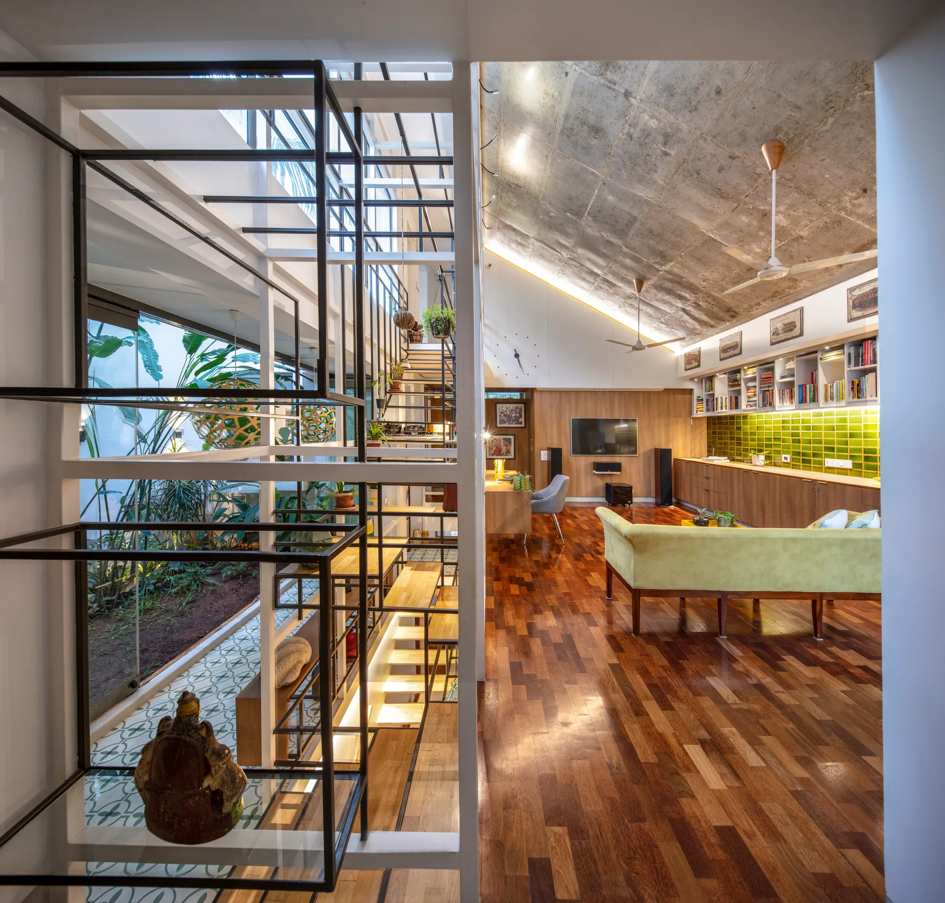A modern living room with polished wood floors, a sloped concrete ceiling, and a glass-enclosed indoor atrium containing plants and stairs.