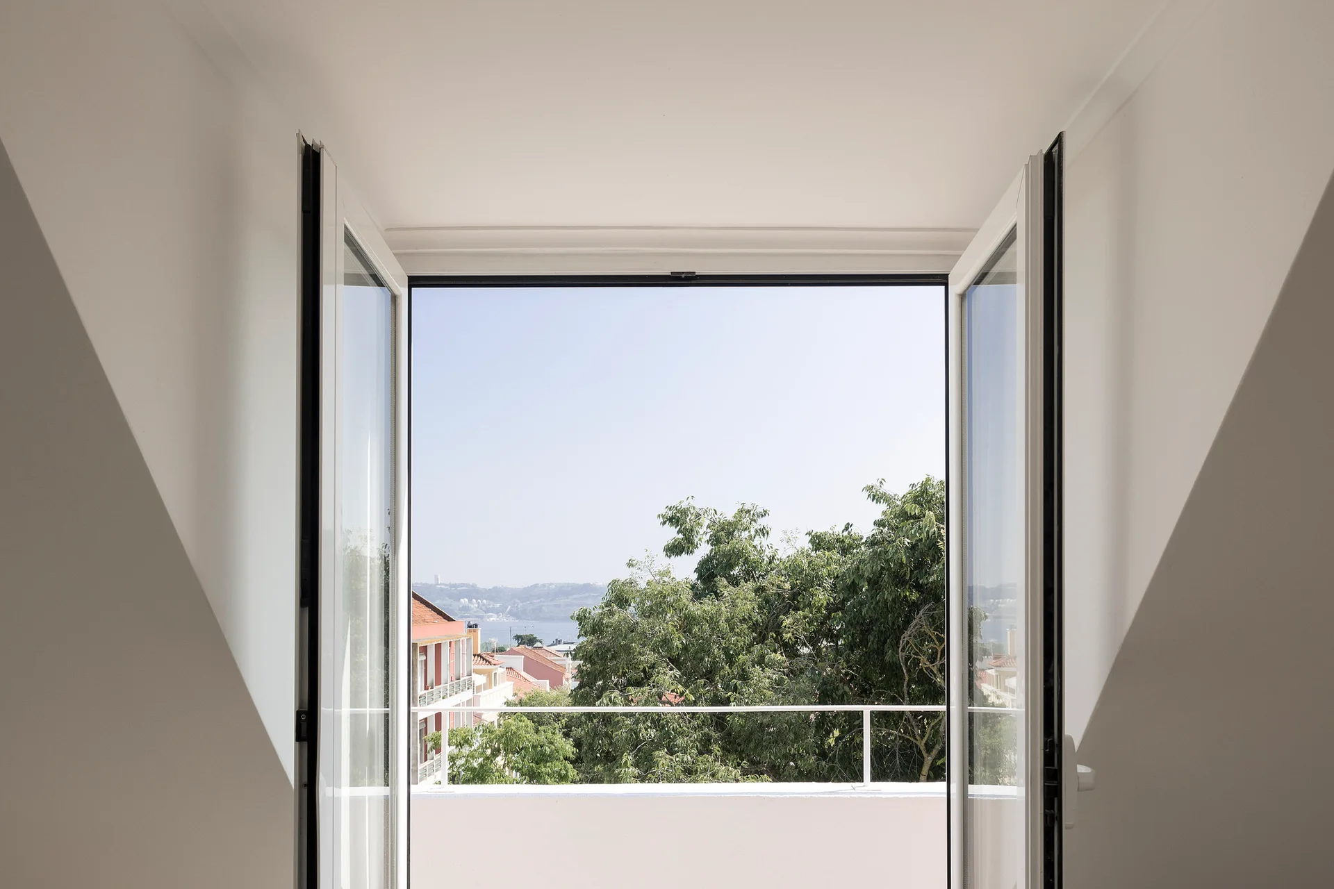 Open white double doors lead to a small balcony with a white metal railing overlooking green trees and terracotta roofed buildings under a clear sky.