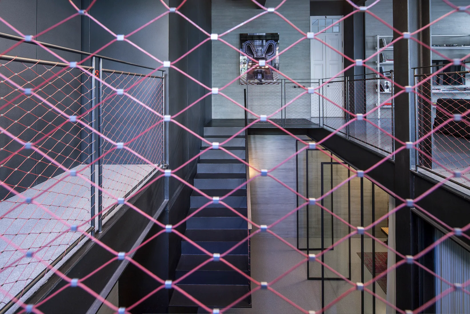A modern multi-level interior features a dark metal staircase and board-formed concrete walls viewed through a red diamond-mesh safety net.