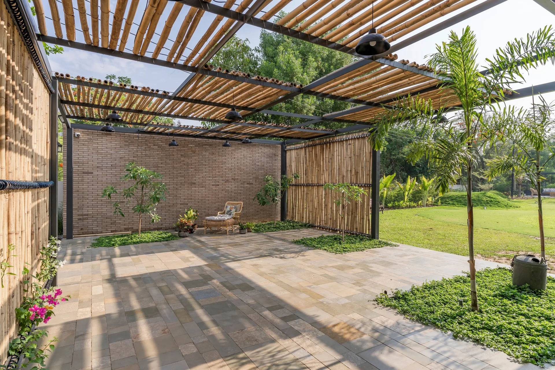 An outdoor patio features a black metal pergola frame with bamboo slatted roofing, stone pavers, vertical bamboo privacy screens, and a brick wall.