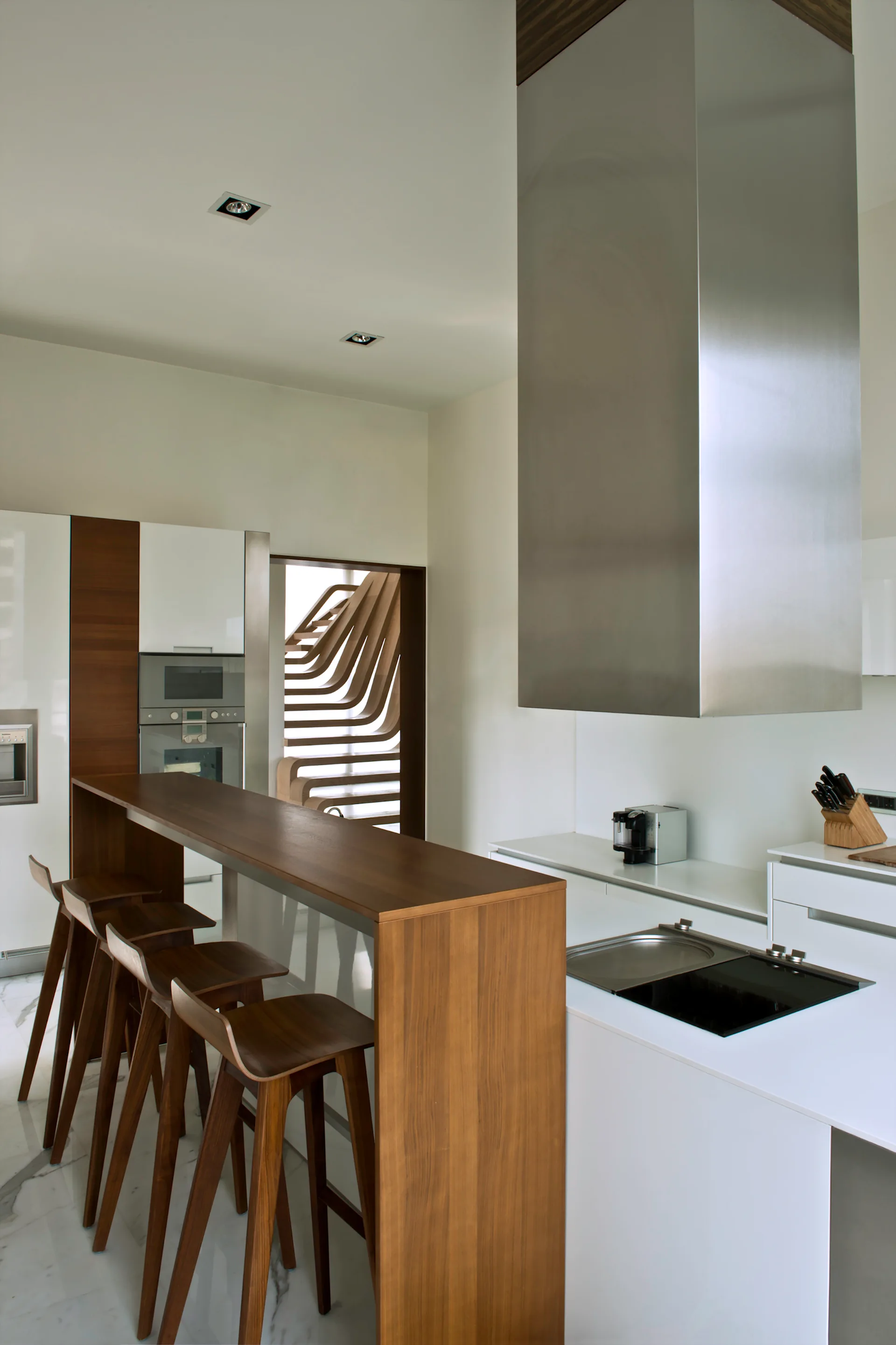 A modern white kitchen features a dark wood breakfast bar with matching stools, a stainless steel range hood, and a wooden staircase in the background.