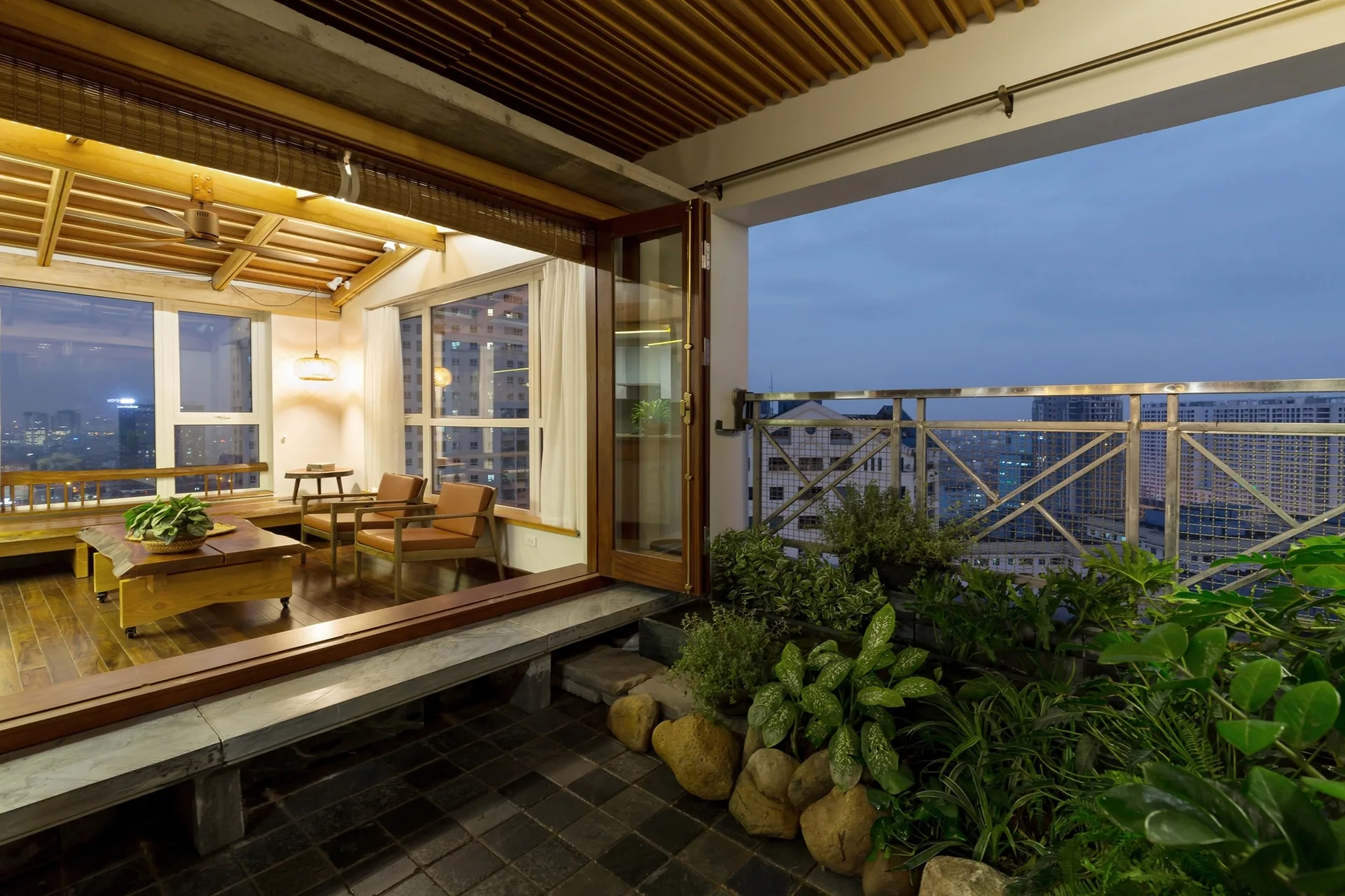 An urban terrace with potted plants and large stones next to an indoor lounge featuring a wood slatted ceiling and tan leather chairs.