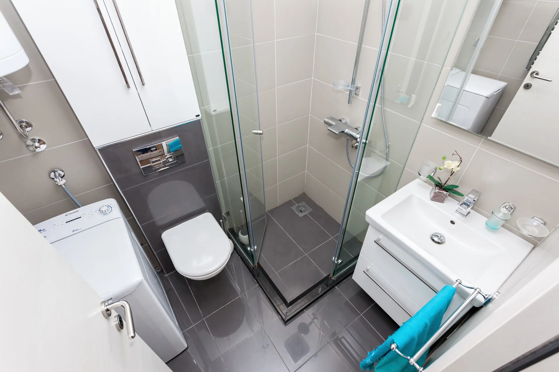A compact grey and white bathroom featuring a glass-walled corner shower, a wall-mounted toilet, a white vanity sink, and a top-loading washing machine.