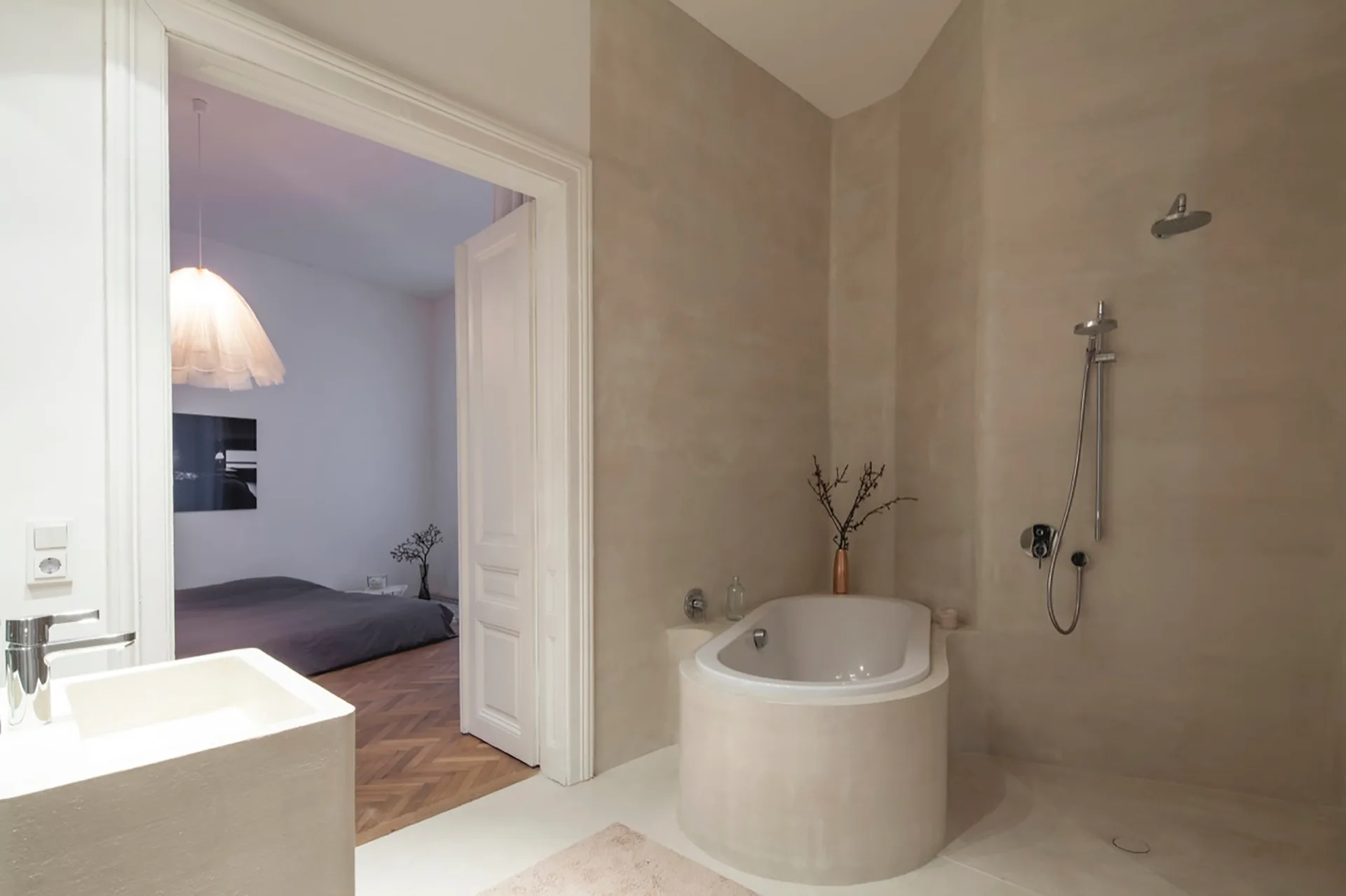 A minimalist beige plaster bathroom featuring an integrated corner bathtub, wall-mounted shower fixtures, and an open doorway leading to a bedroom.