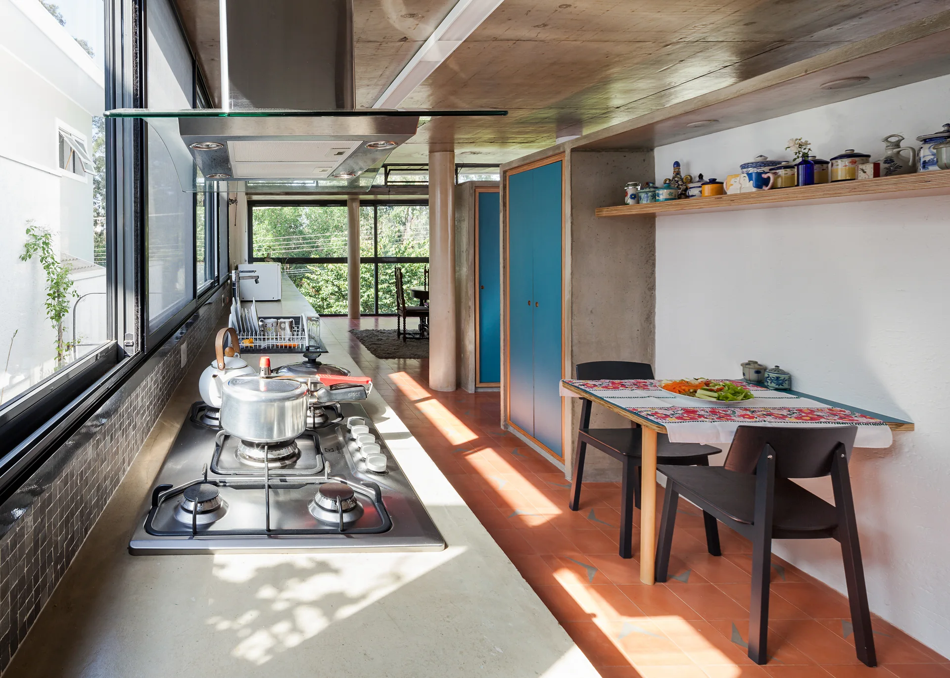 A modern kitchen featuring an exposed concrete ceiling, terracotta tile floor, blue cabinetry, and a small dining table with black chairs.