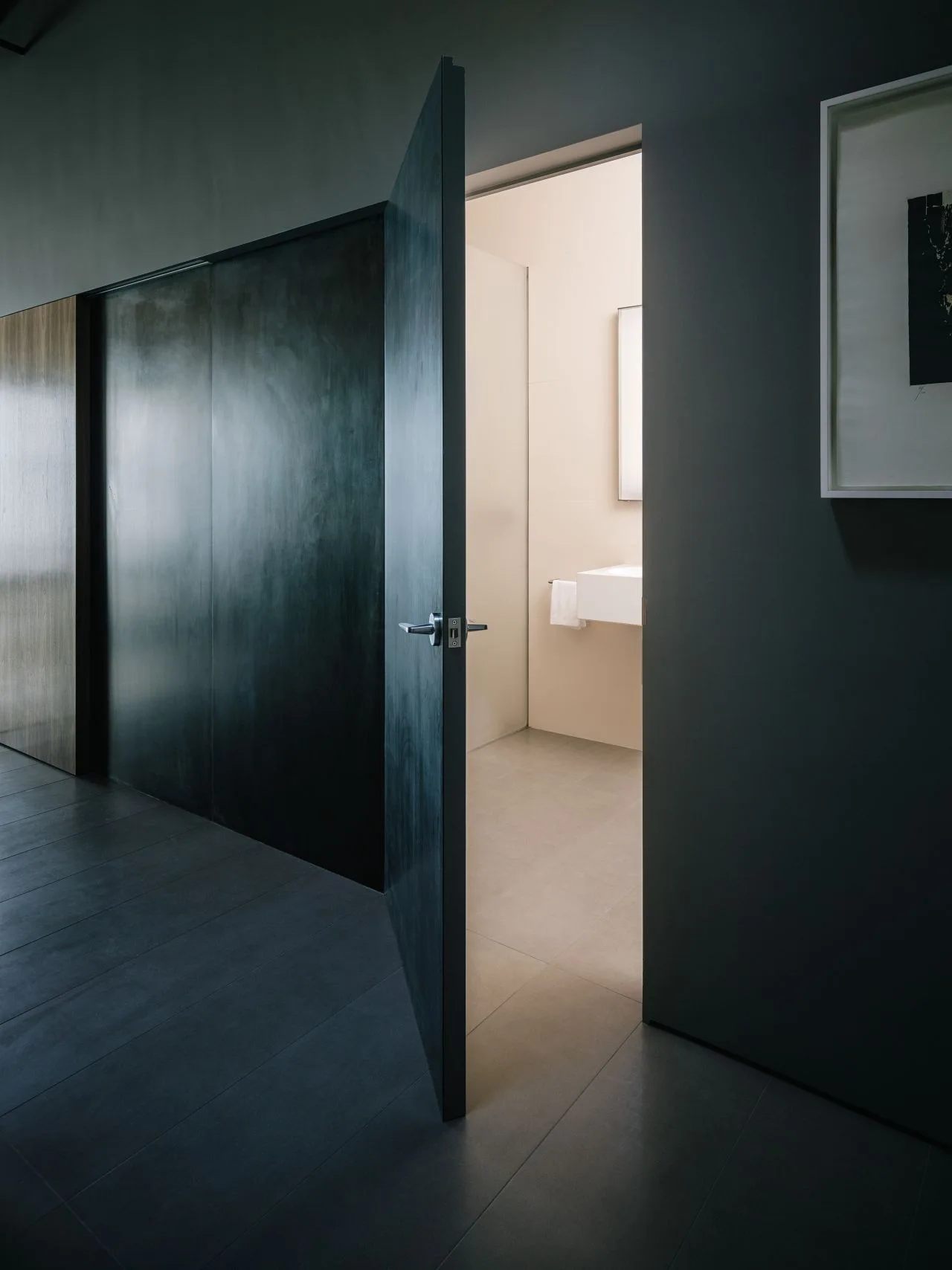 An open dark gray door connects a moody charcoal hallway to a brightly lit minimalist bathroom with a white wall-mounted sink.