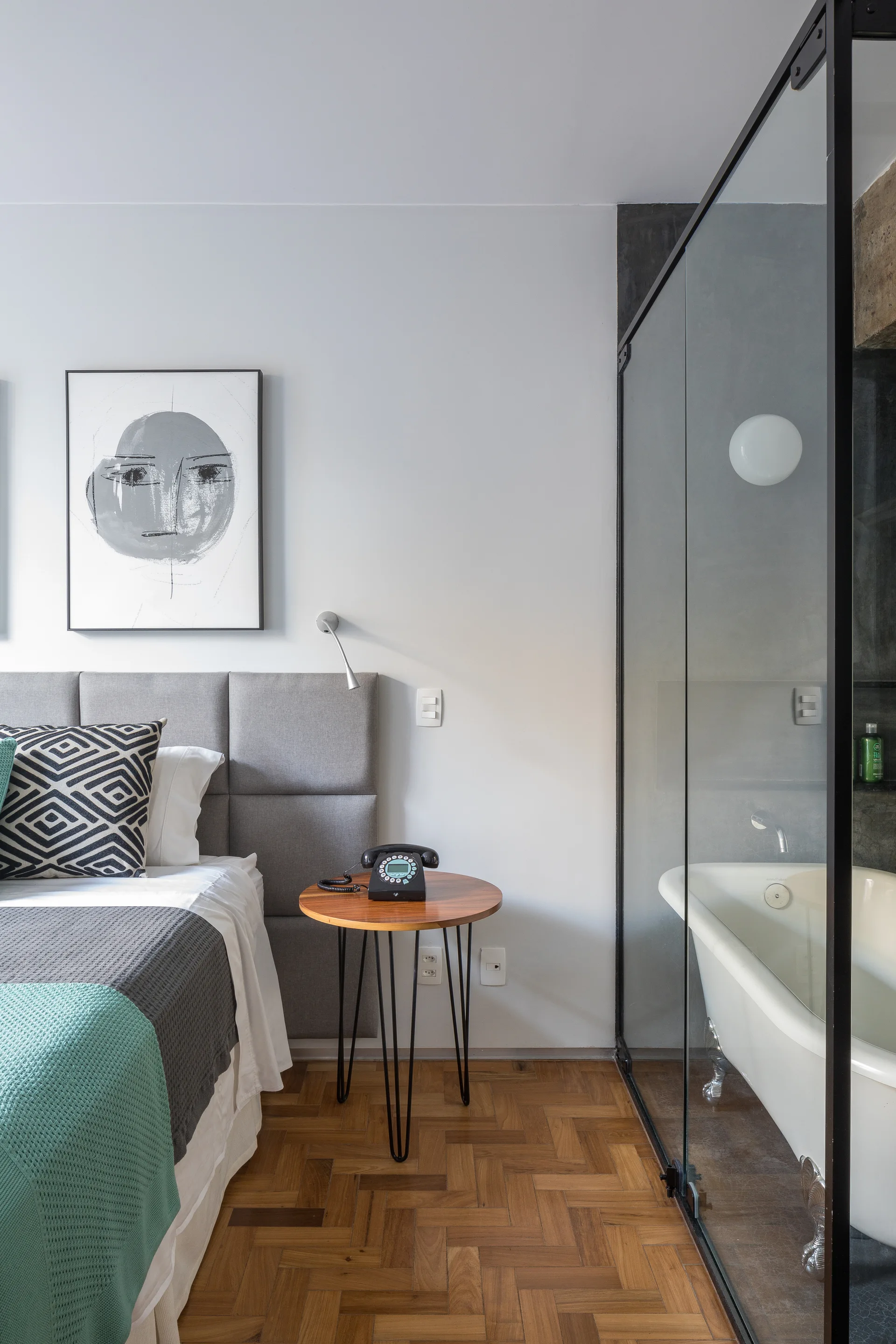 A modern bedroom with a grey upholstered headboard next to a glass-walled bathroom showing a clawfoot bathtub.