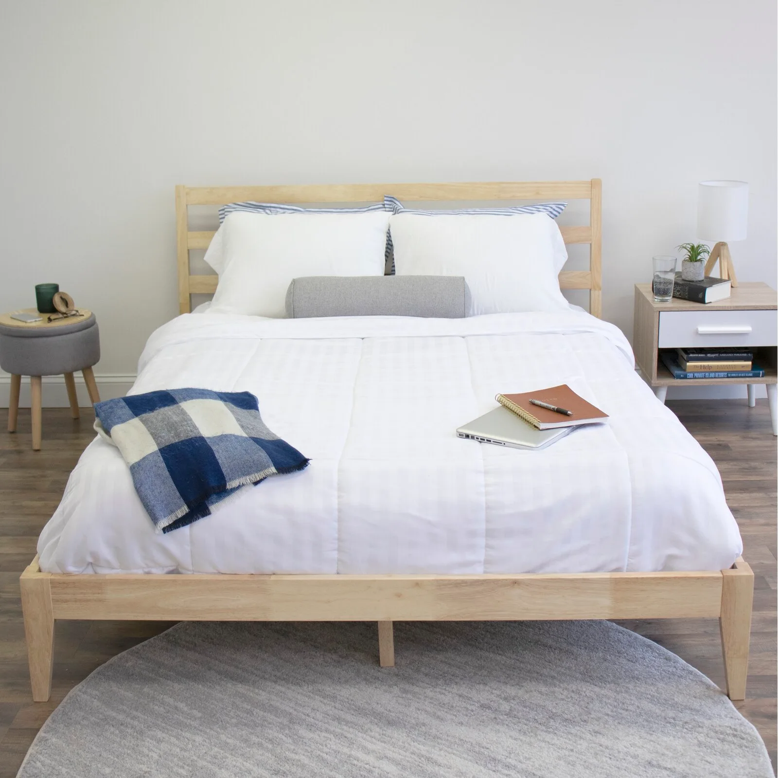 A minimalist bedroom featuring a light wood bed with white bedding, a grey bolster pillow, and a round grey rug on wood floors.