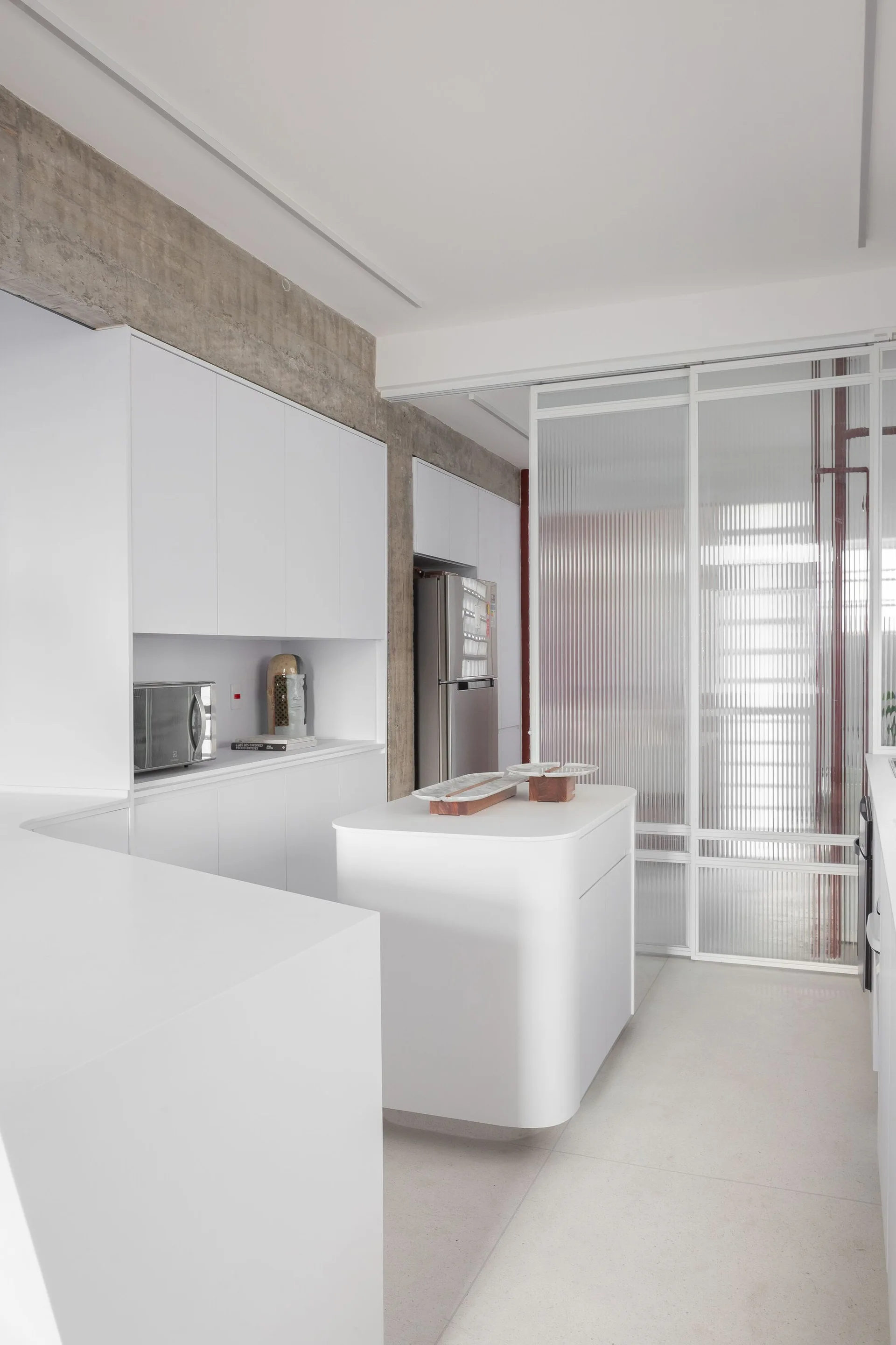A minimalist white kitchen featuring a curved island, flat-front cabinetry, an exposed concrete ceiling beam, and a fluted glass sliding partition.