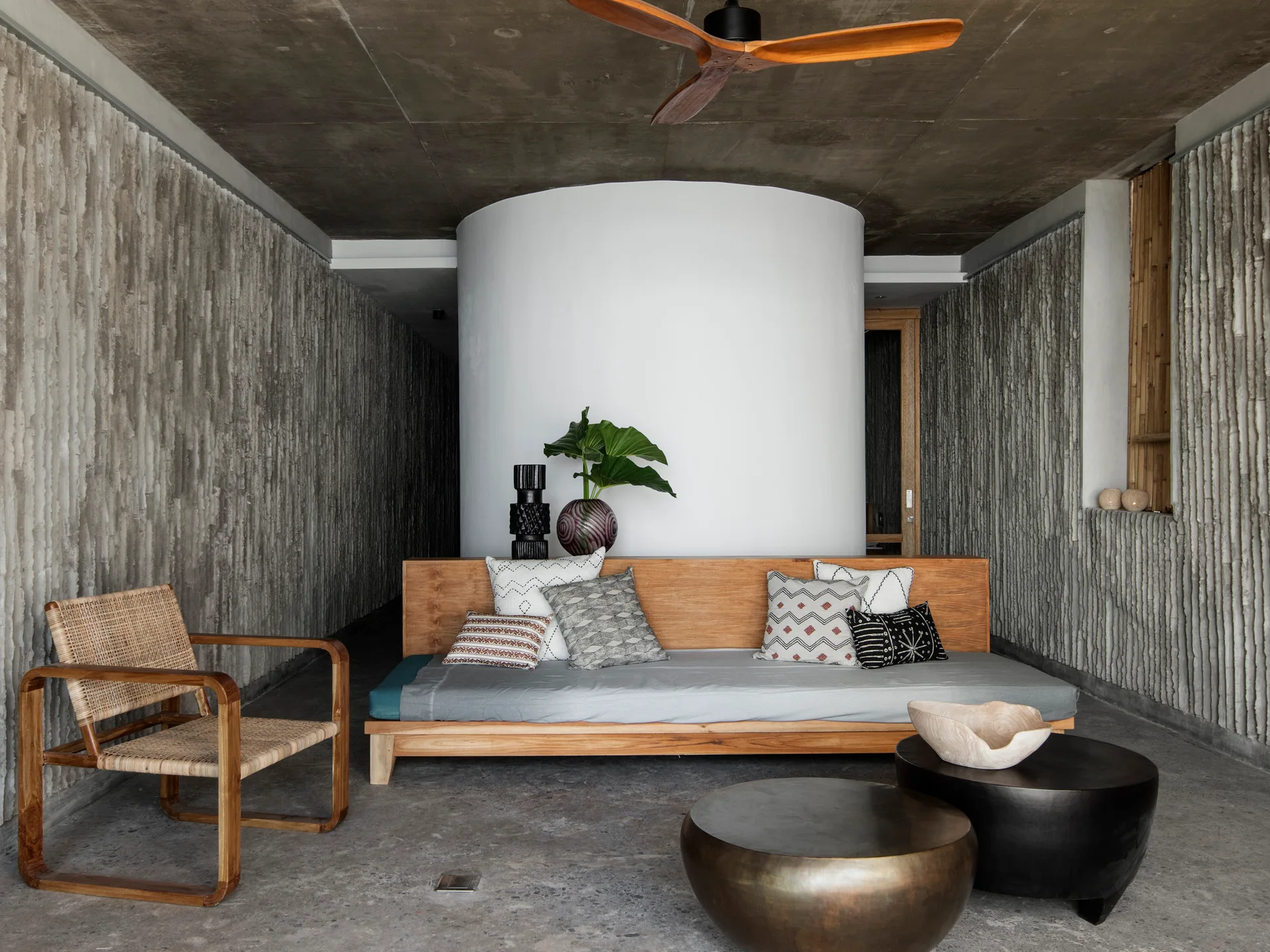 A low wood platform daybed sits in front of a white cylindrical wall, flanked by textured gray concrete walls and a rattan armchair.