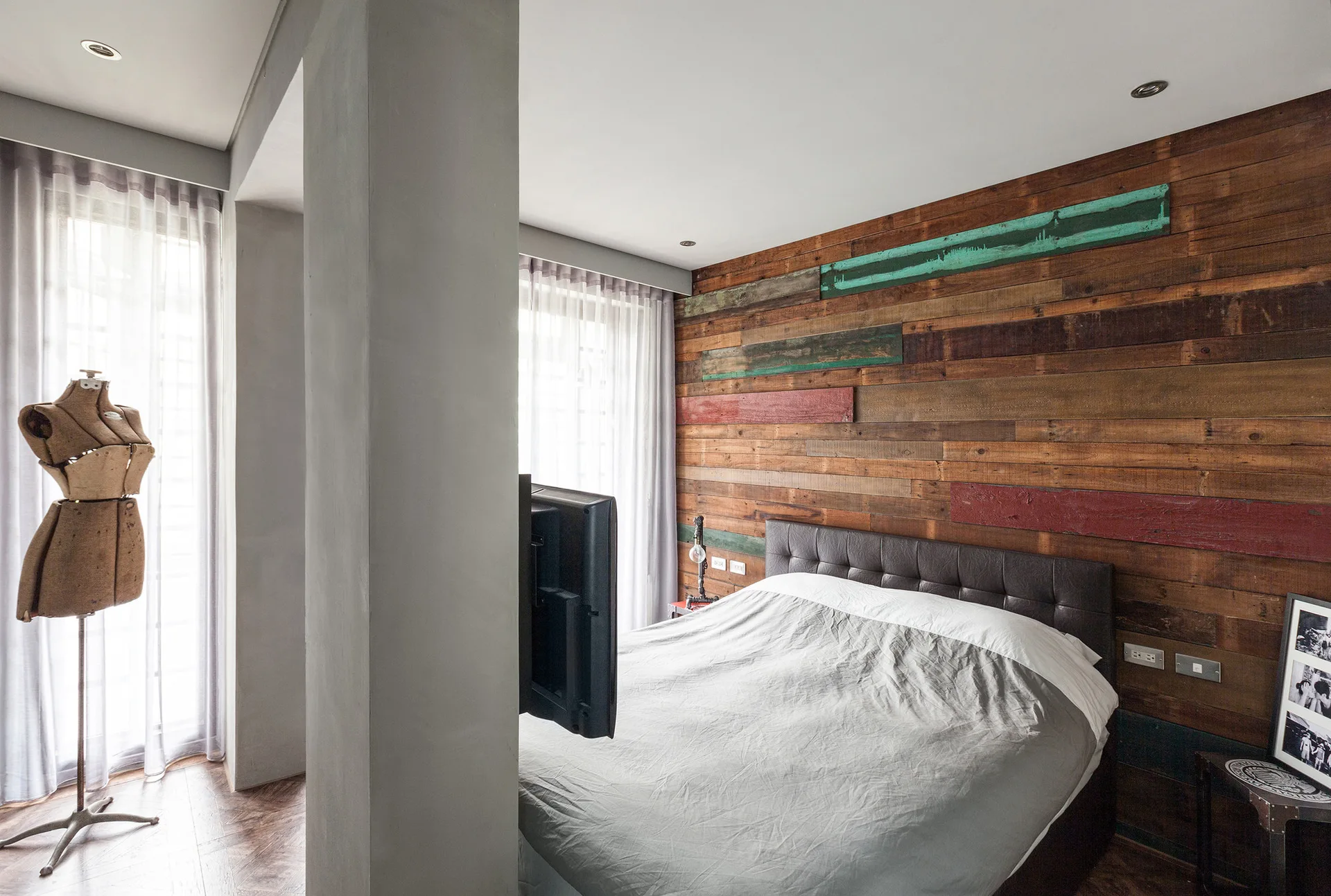 An industrial bedroom features a reclaimed wood accent wall behind a leather bed, with a concrete pillar holding a mounted television.