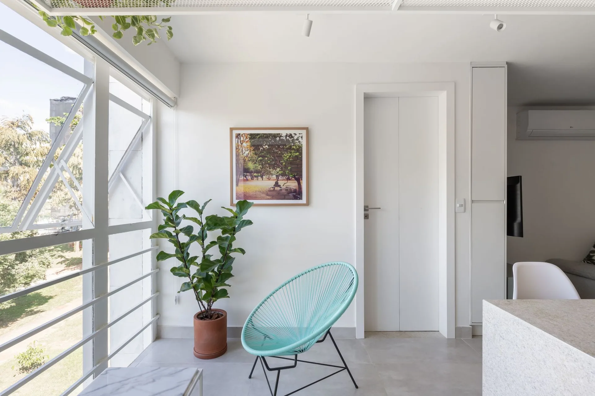 A bright minimalist room features a teal woven Acapulco chair next to a potted plant, a large window, and a white interior door.