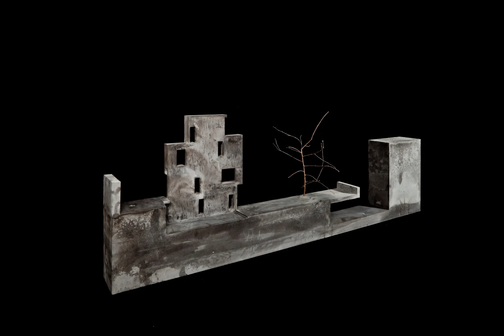 A grey concrete architectural model featuring minimalist geometric structures and a single bare branch, isolated against a solid black background.
