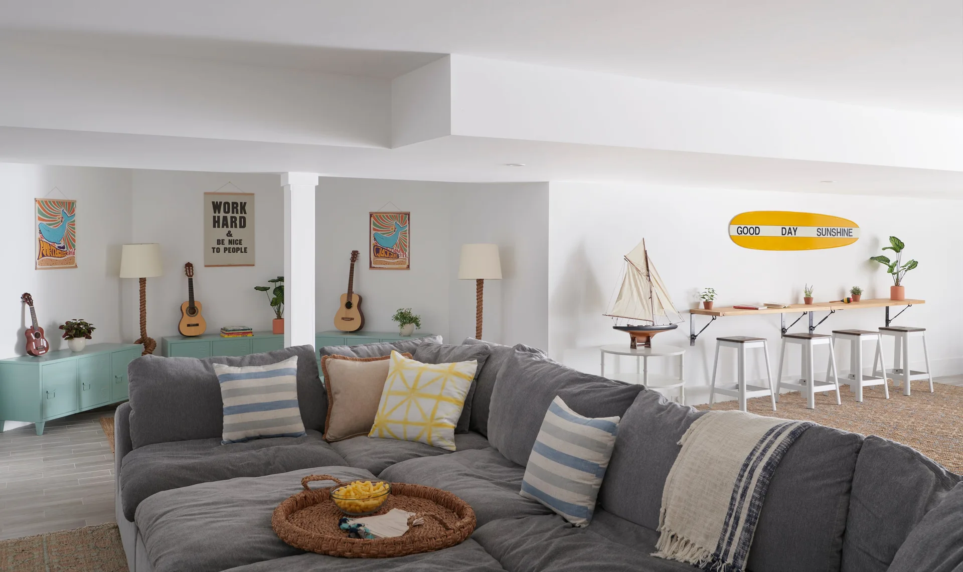 A casual basement living room features a large gray sectional sofa, light blue metal storage cabinets, and a wood-topped bar with white stools.