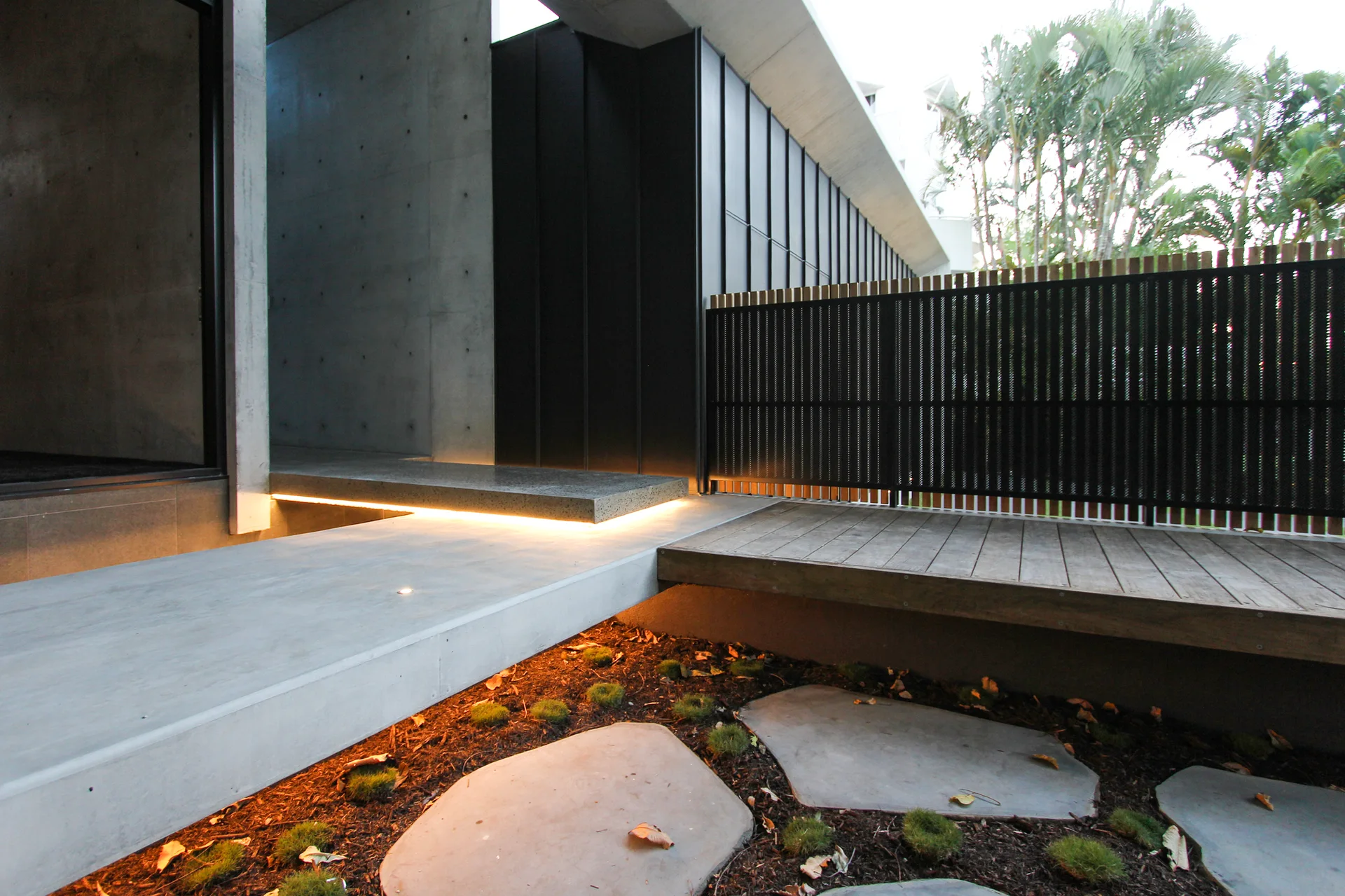 A modern outdoor terrace features a floating concrete walkway, a wooden timber deck, and a dark slatted fence lit by warm under-step lighting.