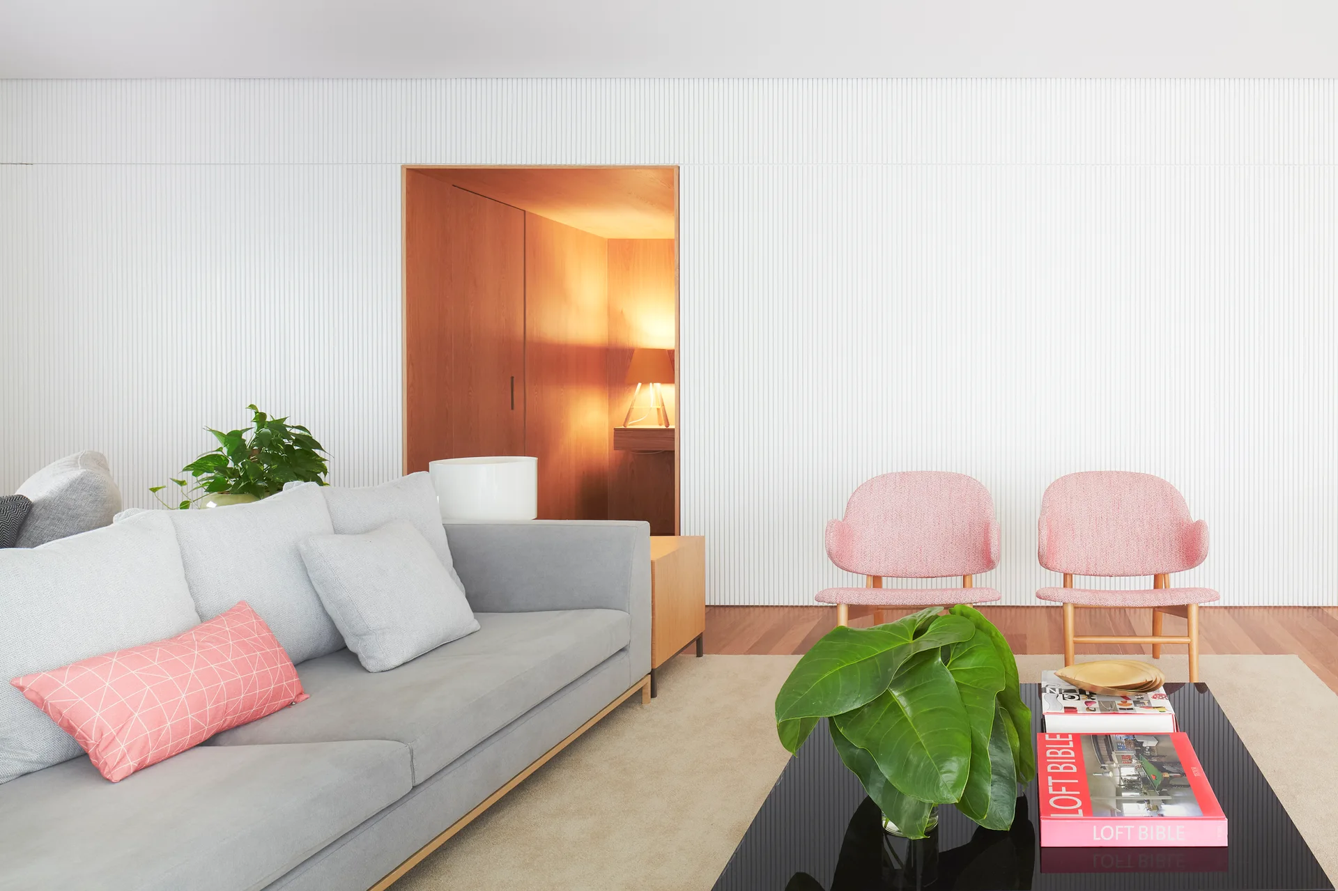 A modern living room features a grey sofa, two pink armchairs, and a white ribbed wall with a recessed wood-lined entryway.