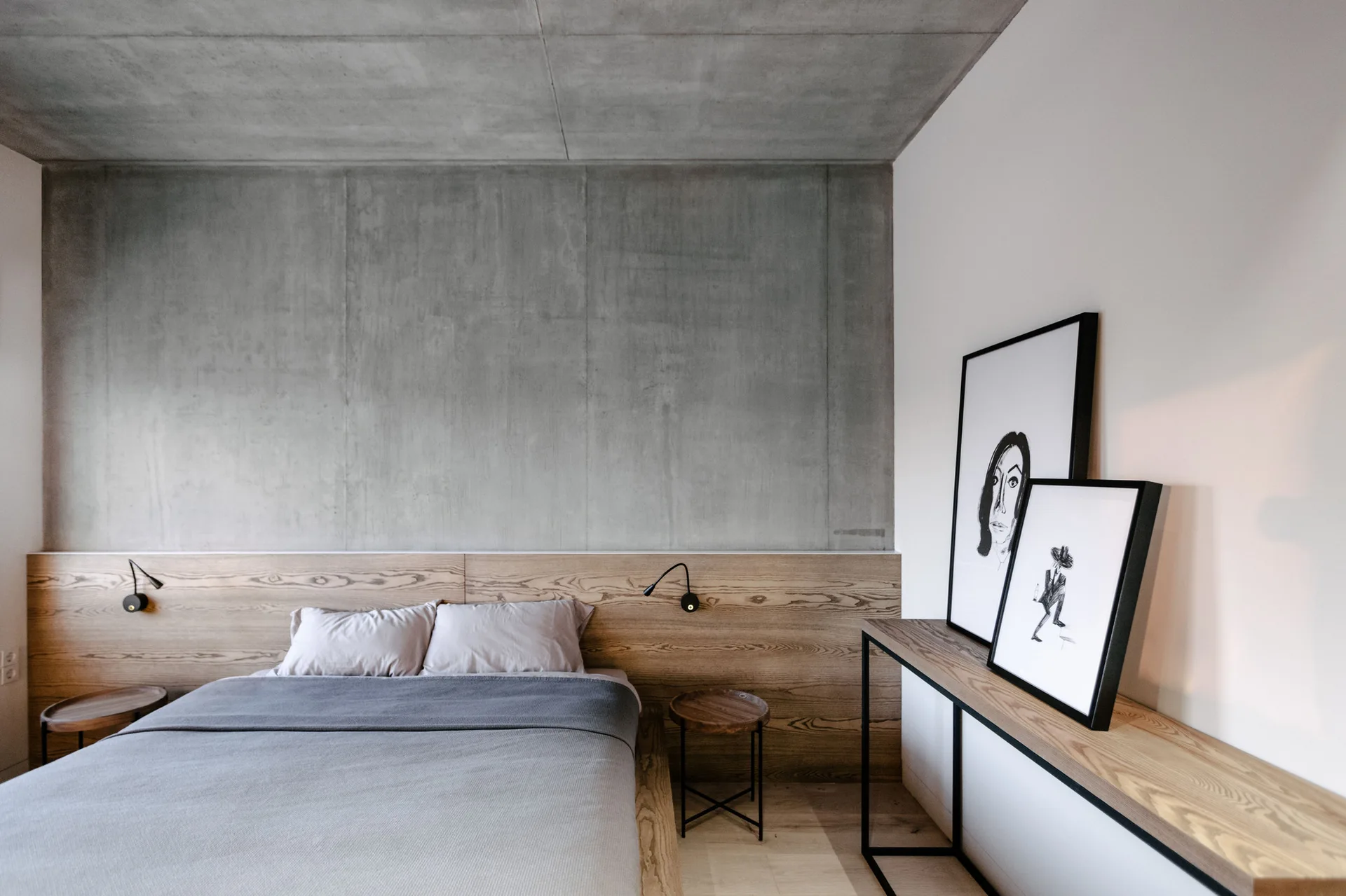 A modern bedroom features a bed with grey bedding set against a low wood headboard beneath an exposed concrete wall and ceiling.