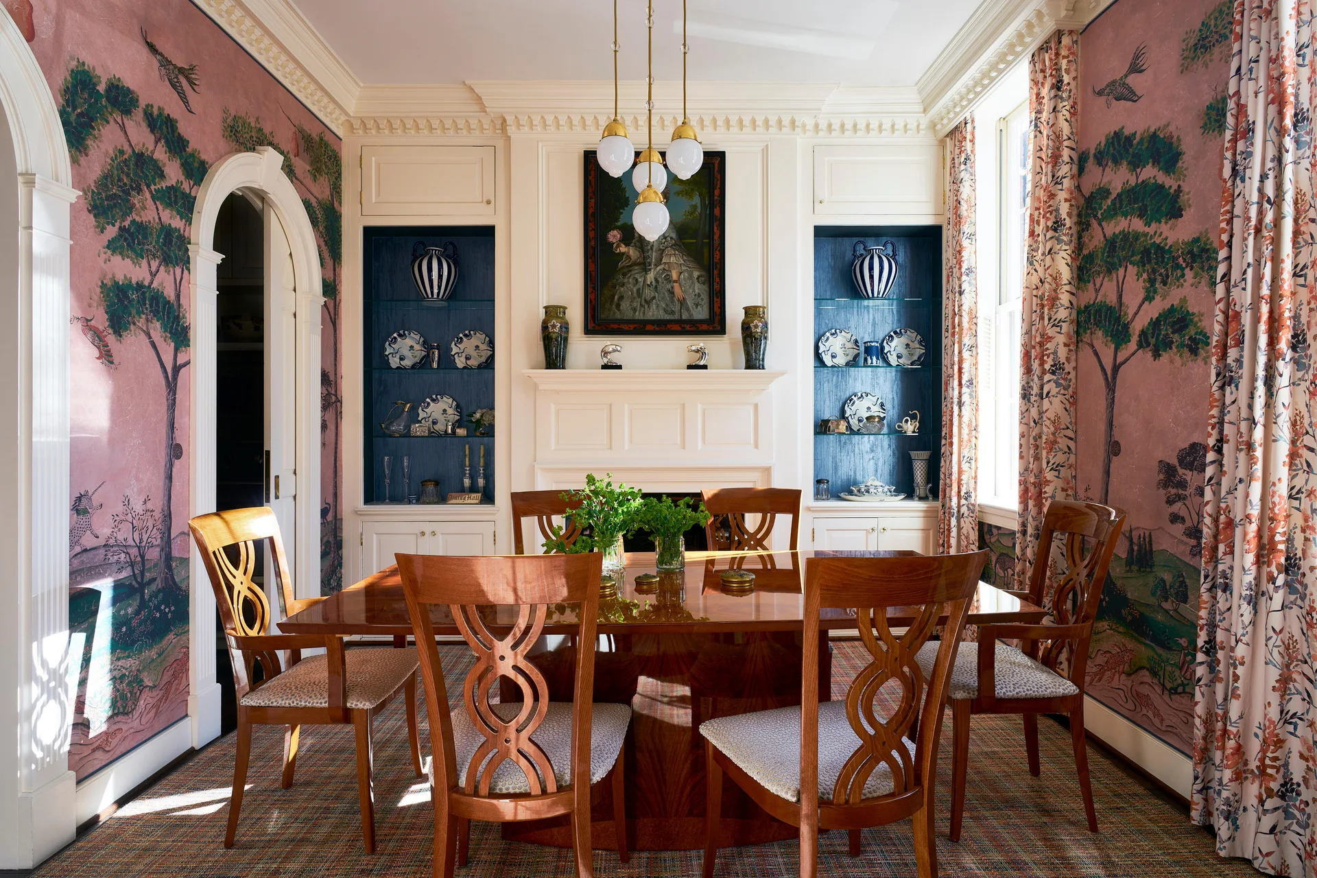 A traditional dining room featuring a polished wood table, pink scenic wallpaper, and a white fireplace flanked by blue-backed built-ins.