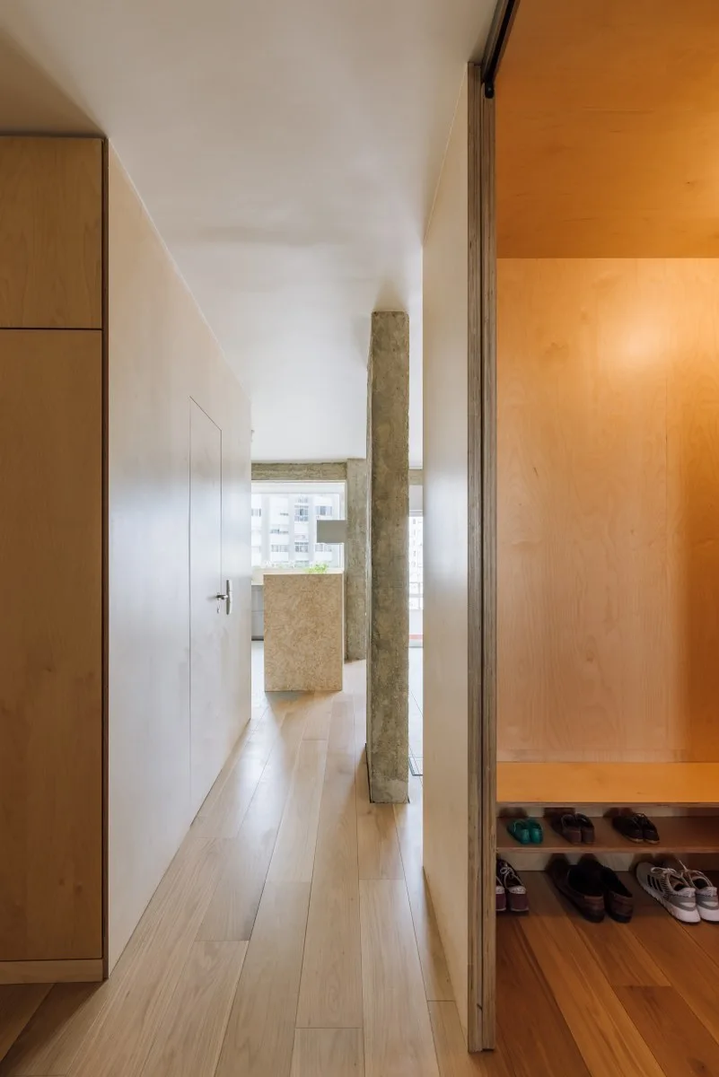 A minimalist hallway features light plywood paneled walls with a flush door, an exposed concrete pillar, and a built-in wooden entryway shoe bench.